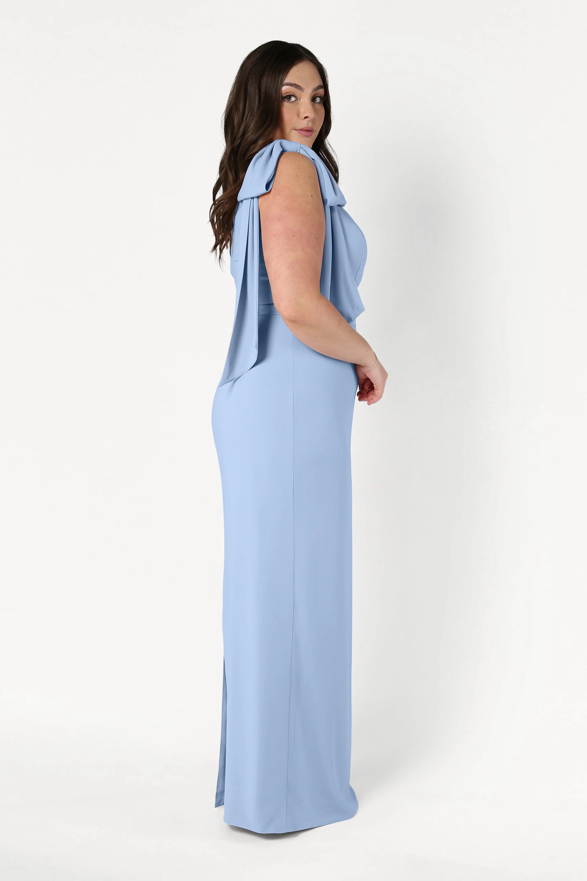A woman with long, dark hair stands in profile wearing the TH&TH Valentina Bridesmaid Dress in Dusty Blue, featuring draped sleeves and a size-inclusive fit, set against a plain white background.