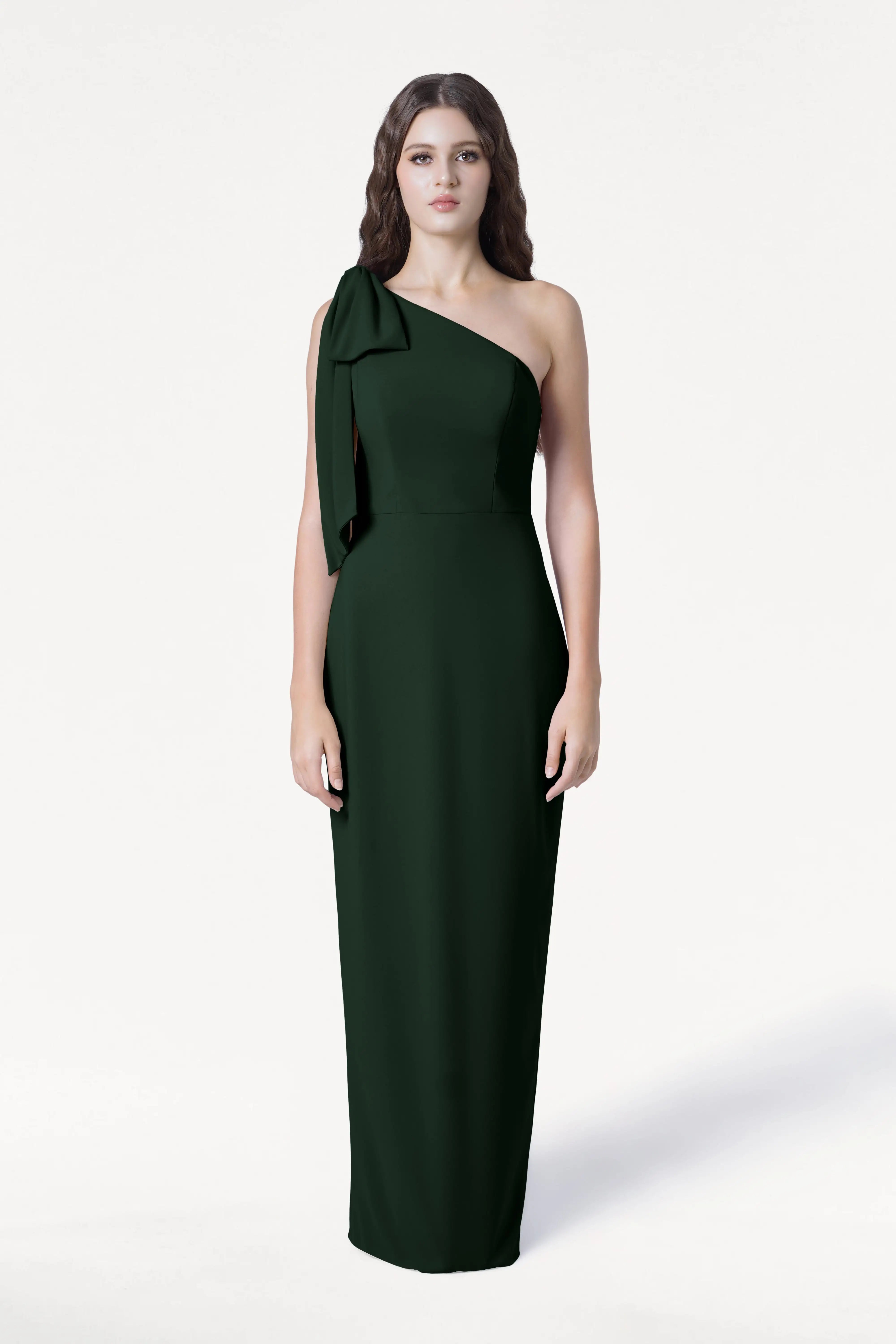 A woman poses against a white background wearing the TH&TH Valentina Bridesmaid Dress in Forest Green, a modern crepe, floor-length, one-shoulder gown with a bow detail. She has long brown hair and a neutral expression.