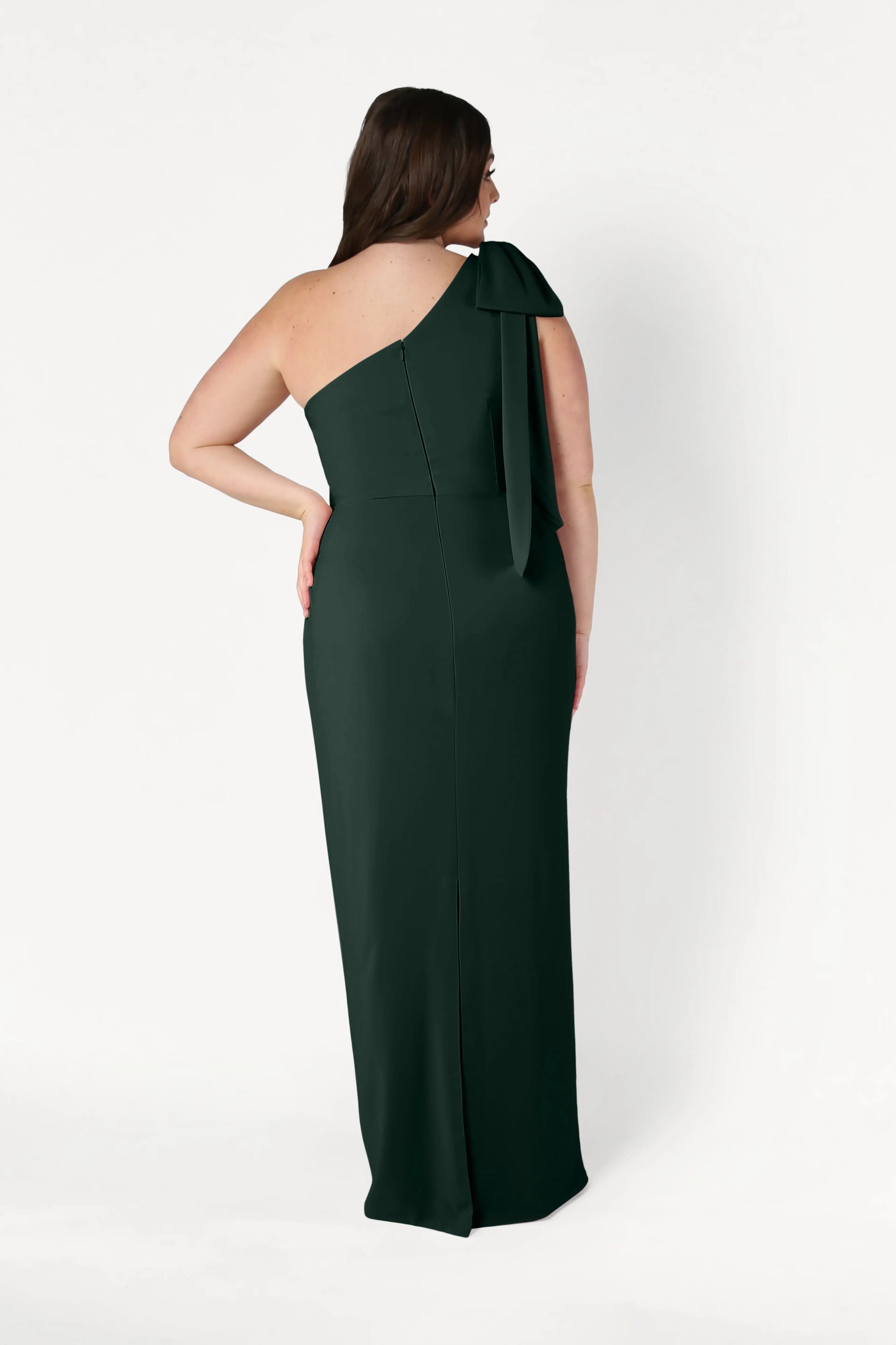 A woman faces away from the camera in a TH&TH Valentina Bridesmaid Dress in Forest Green—a modern, floor-length crepe gown with a one-shoulder bow—set against a plain white background.