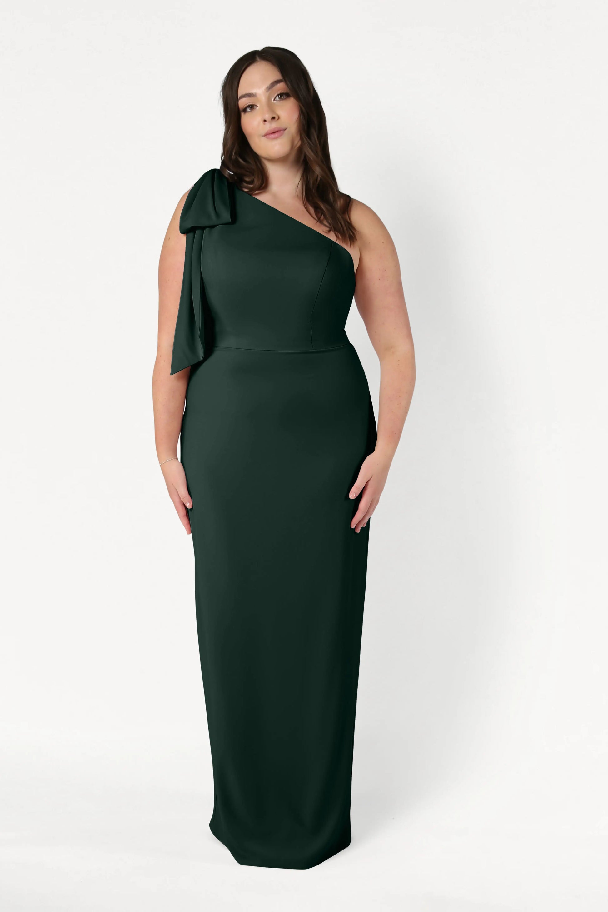 A woman wears the TH&TH Valentina Bridesmaid Dress in Forest Green, featuring a one-shoulder design with a bow detail, standing against a plain white background.