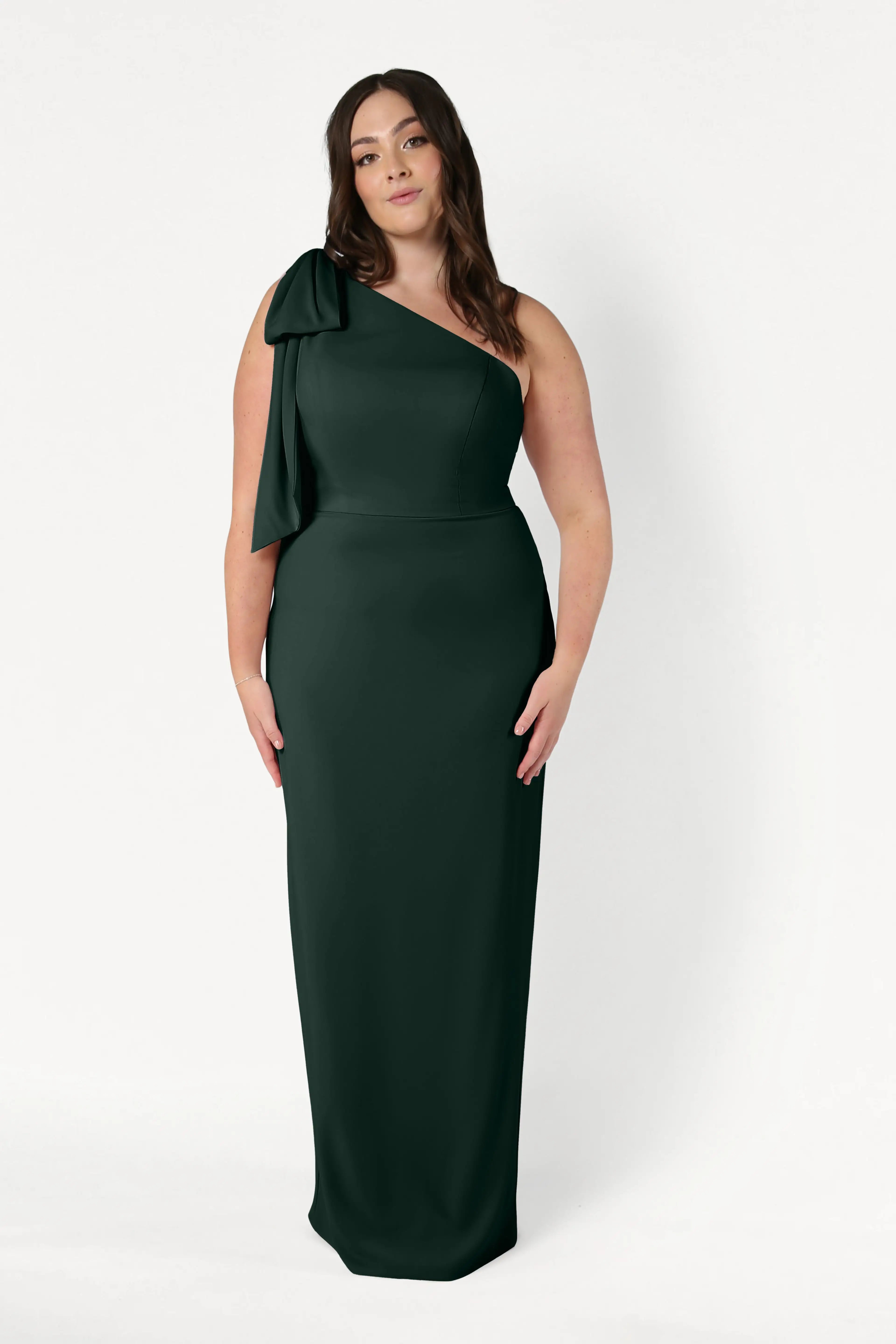 A woman wears the TH&TH Valentina Bridesmaid Dress in Forest Green, featuring a one-shoulder design with a bow detail, standing against a plain white background.
