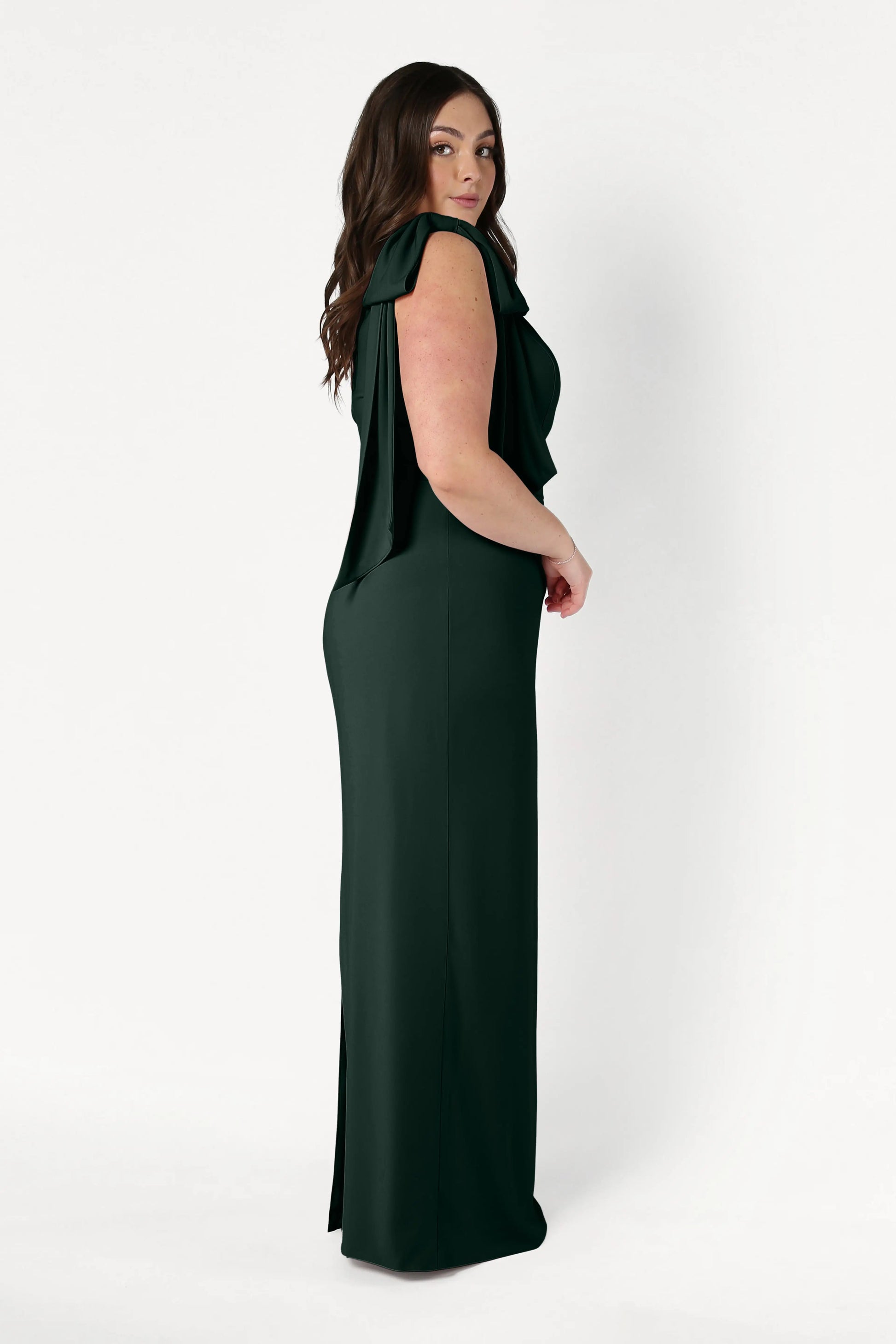 A woman with long brown hair stands sideways in the TH&TH Valentina Bridesmaid Dress in Forest Green, a size-inclusive, floor-length crepe gown featuring draped shoulder detailing, set against a plain white background.