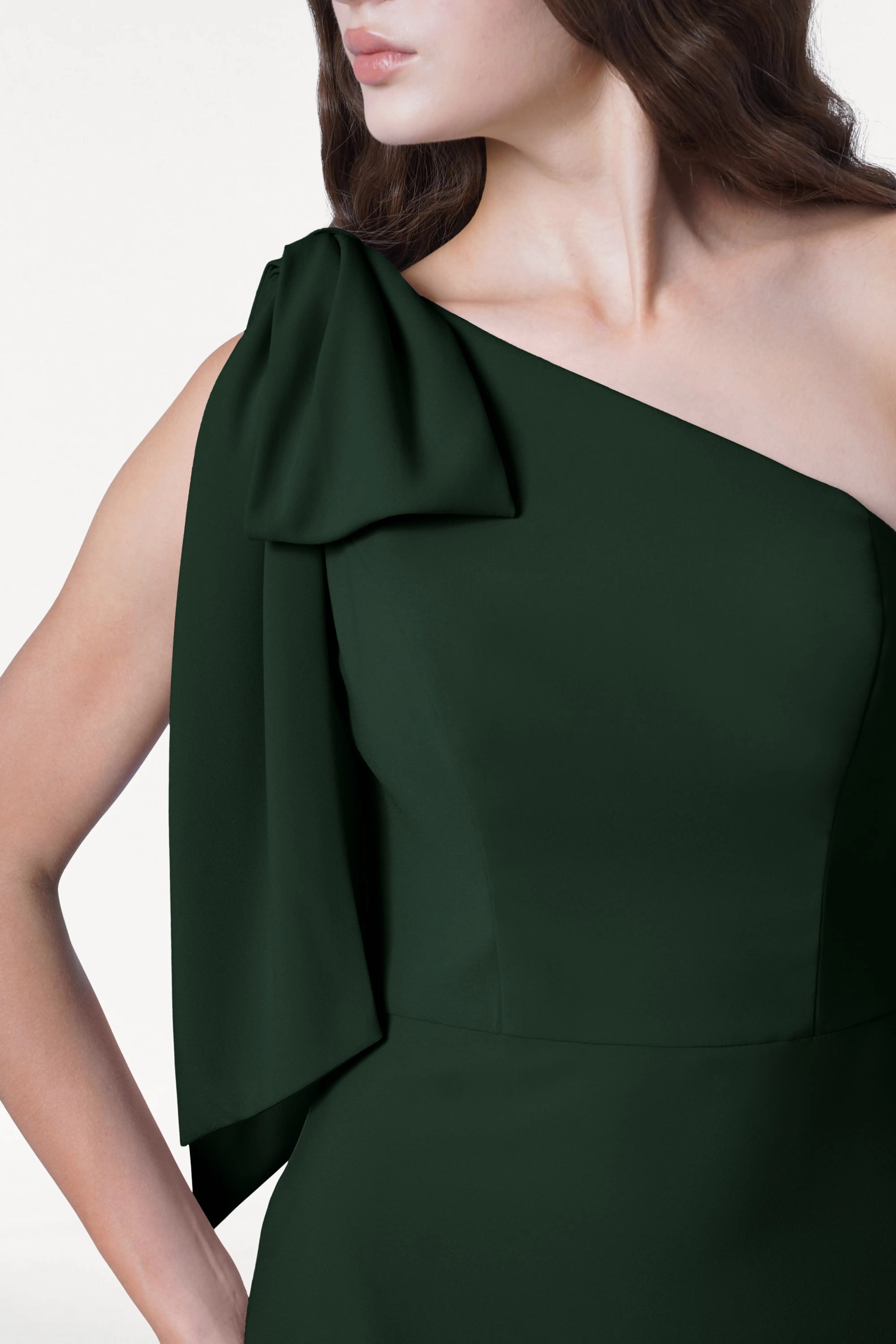 A woman models the TH&TH Valentina Bridesmaid Dress in Forest Green—a modern crepe one-shoulder style with a draped sleeve—against a white background. Her brown hair is loose, and her face is partly out of frame.