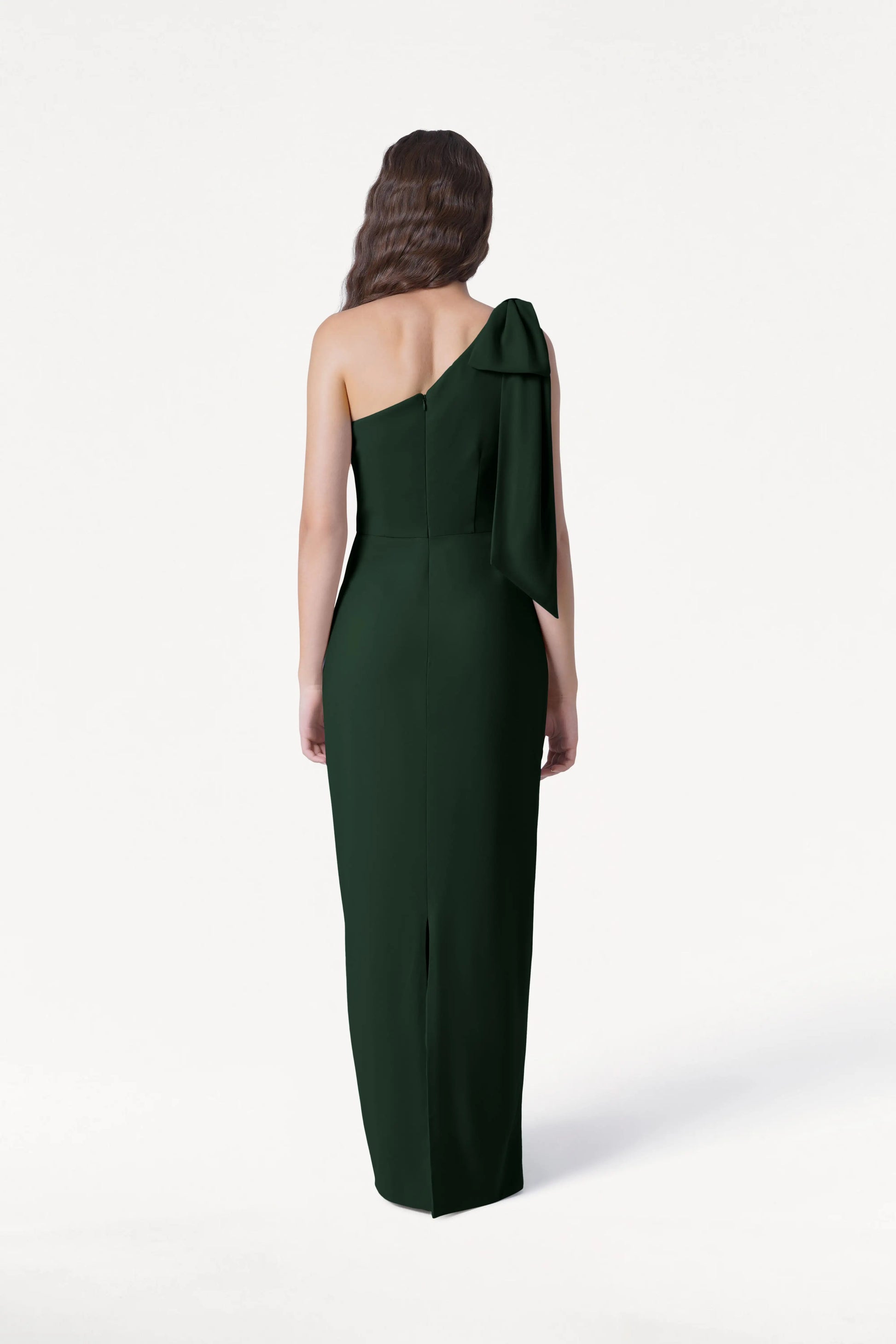 A woman with long wavy brown hair faces away, wearing the size-inclusive Valentina Bridesmaid Dress in Forest Green by TH&TH—a modern, one-shoulder style with a large bow—set against a plain white background.