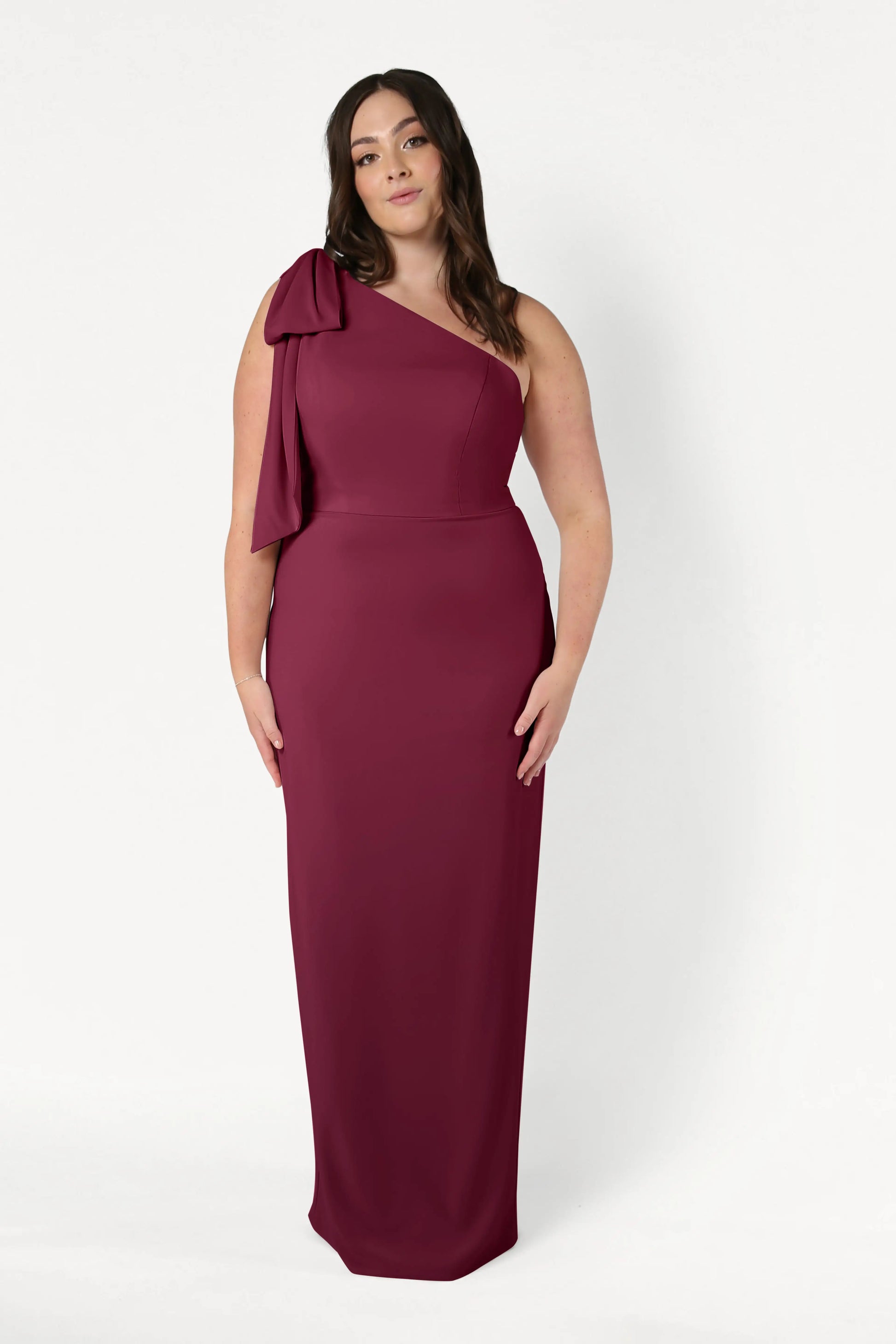 A woman stands against a plain white background wearing the TH&TH Valentina Bridesmaid Dress in Merlot, a sleeveless, floor-length, one-shoulder gown featuring a large bow.