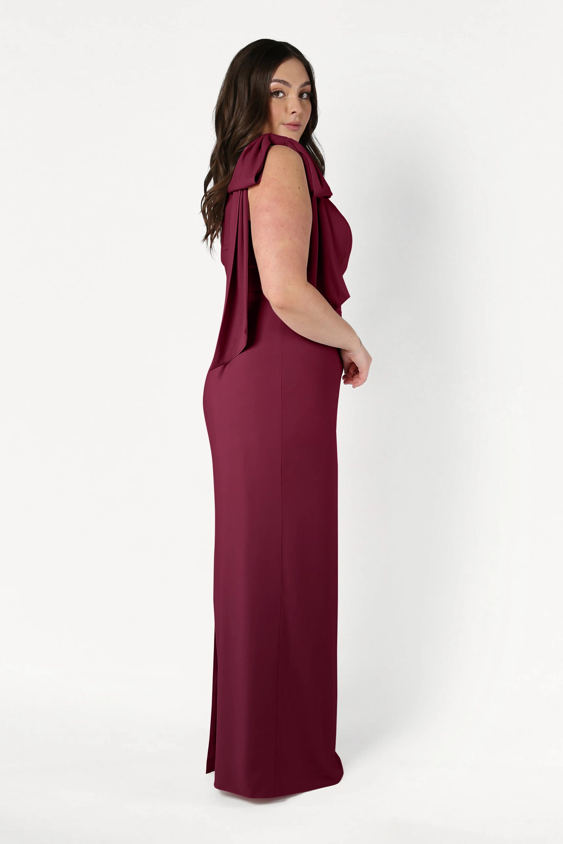A woman with long dark hair stands in profile, wearing the floor-length Valentina Bridesmaid Dress in Merlot by TH&TH, featuring modern crepe fabric and draped shoulders, set against a plain white background.
