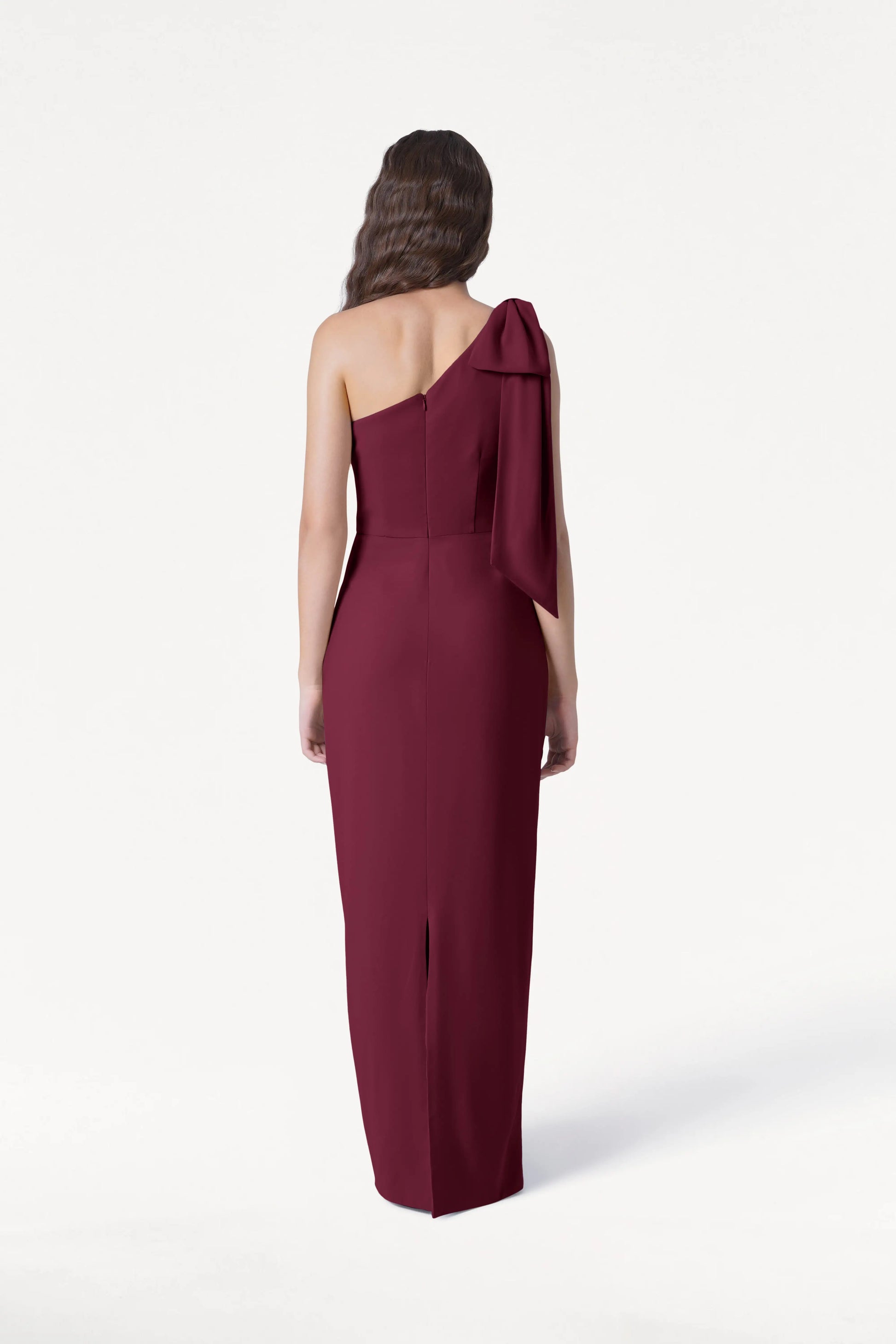 A person with long wavy hair models the TH&TH Valentina Bridesmaid Dress in Merlot, a floor-length one-shoulder gown with a draped bow detail, shown from the back against a plain white background.