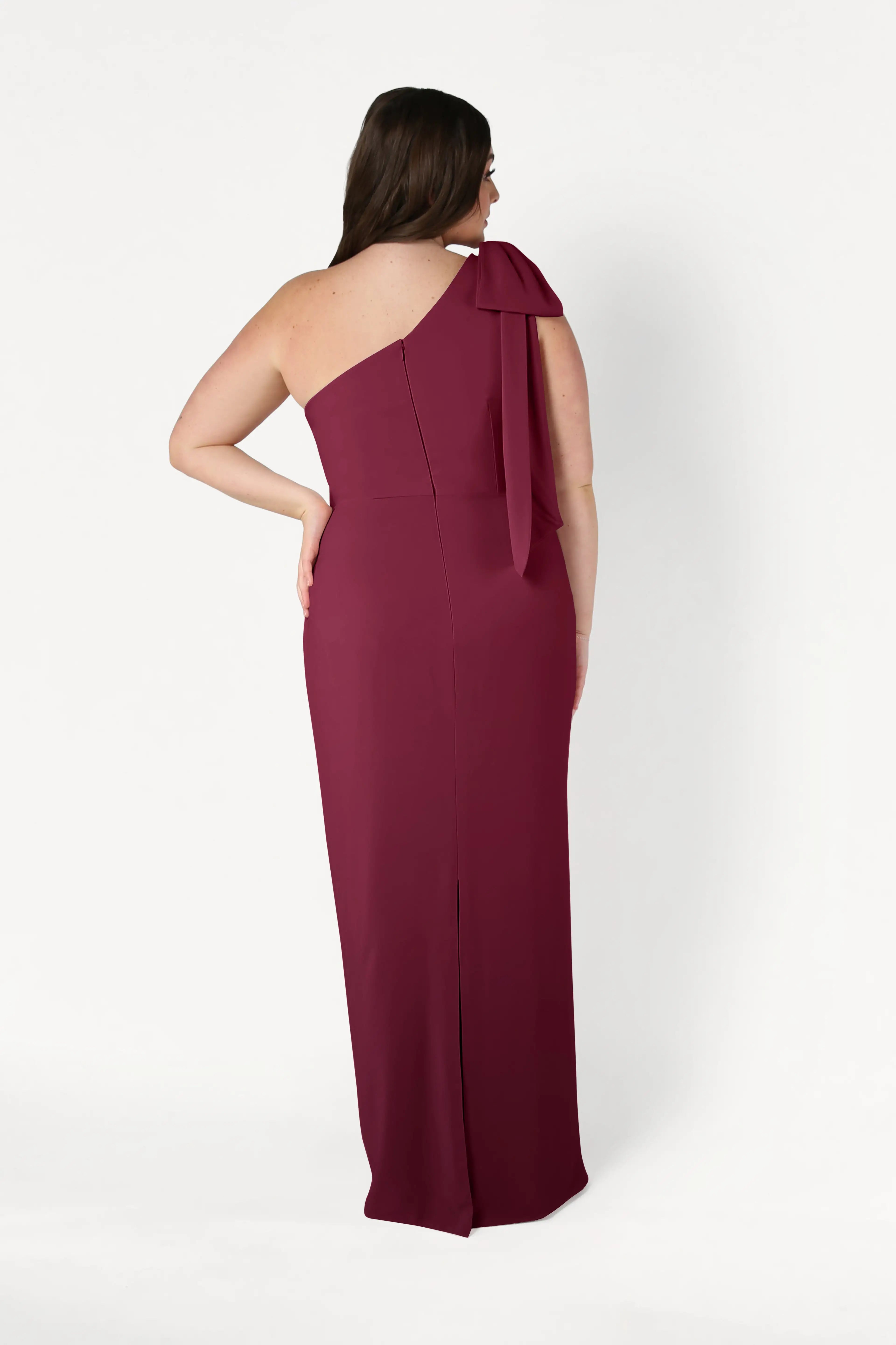A woman faces away, showcasing the elegant back of the TH&TH Valentina Bridesmaid Dress in Merlot—a modern, floor-length, one-shoulder crepe gown with a bow detail—against a plain white background.