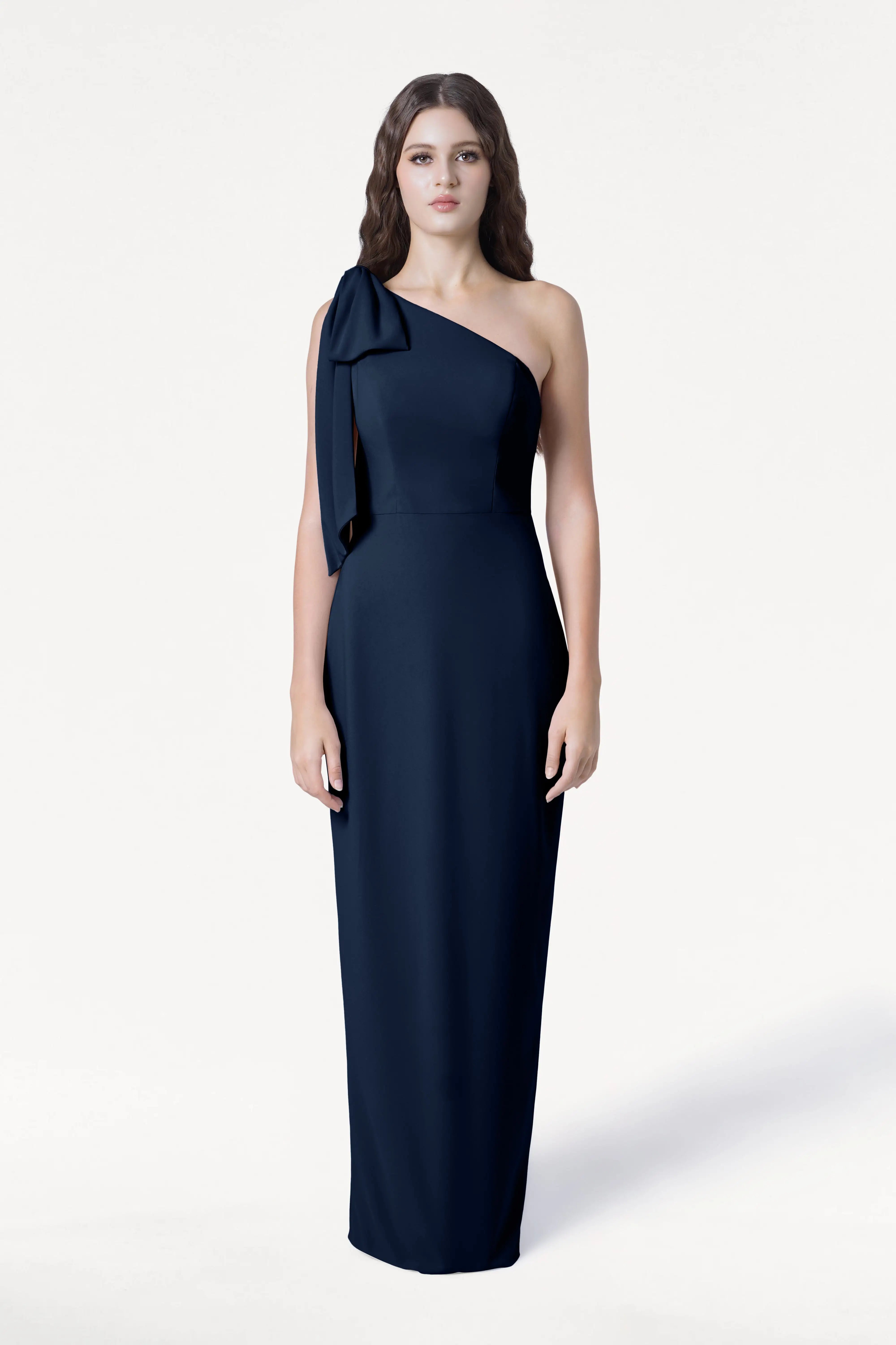 A woman with long, wavy brown hair faces forward against a white background, wearing the TH&TH Valentina Bridesmaid Dress in Navy—a floor-length, one-shoulder crepe gown with a bow on the left shoulder and a neutral expression.
