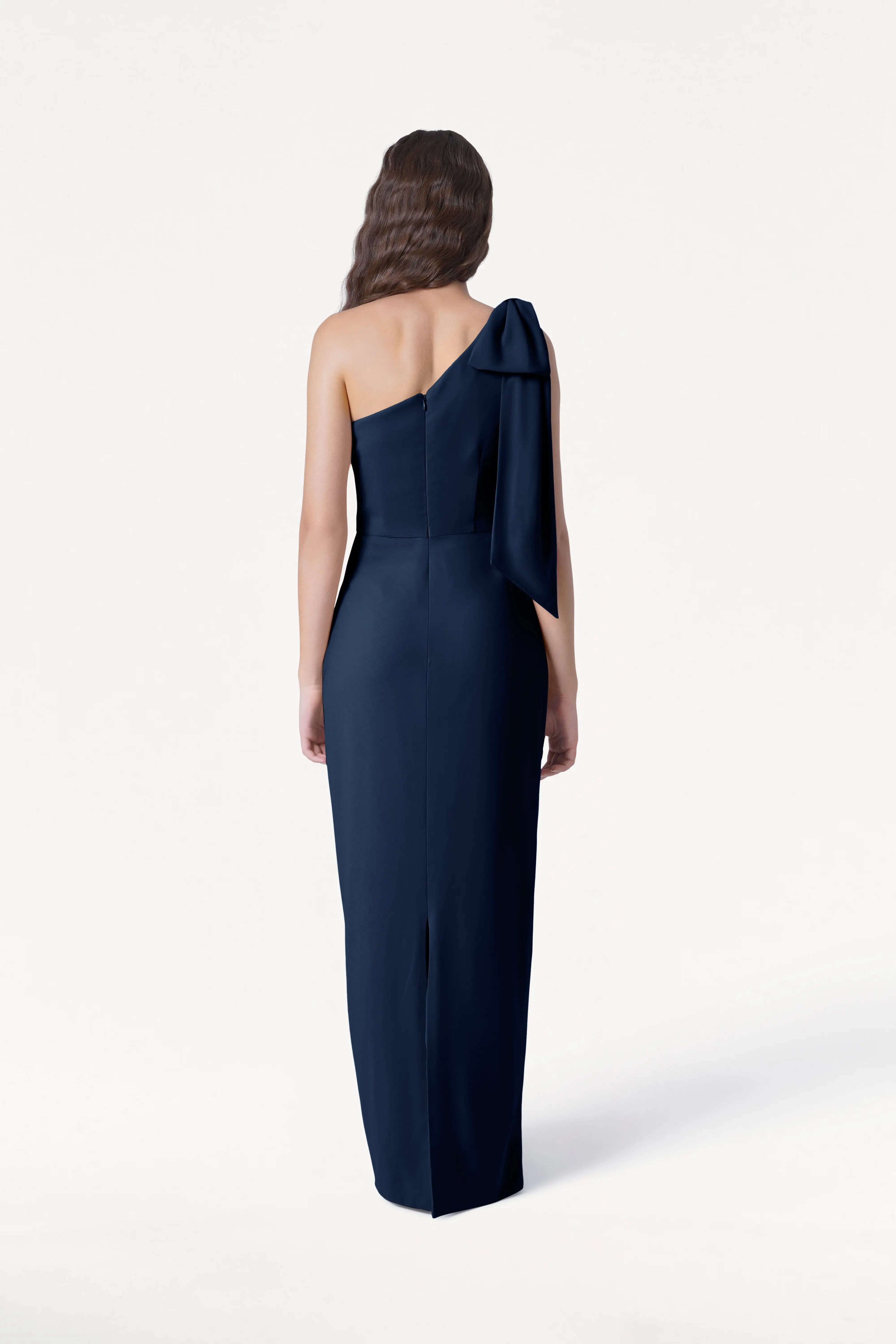 A woman with long wavy hair, seen from behind, wears the TH&TH Valentina Bridesmaid Dress in Navy—a floor-length crepe gown with a large bow on the right shoulder—against a plain white background.