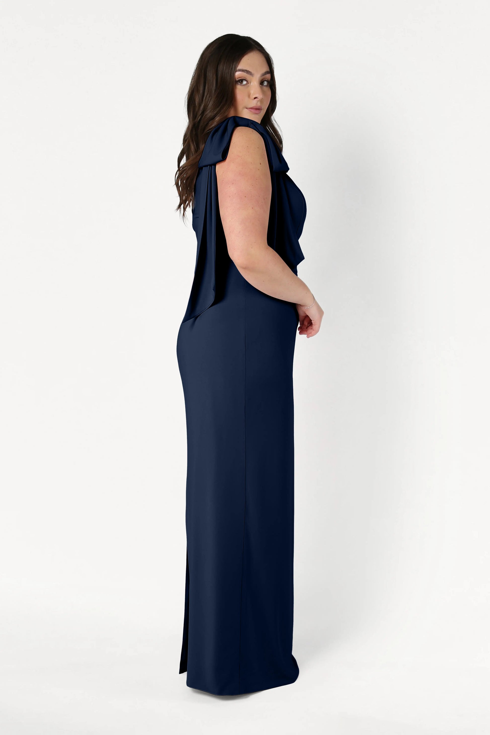 A woman with long brown hair stands sideways against a plain white background, wearing the TH&TH Valentina Bridesmaid Dress in Navy—a floor-length, sleeveless crepe gown with draped shoulder detailing.