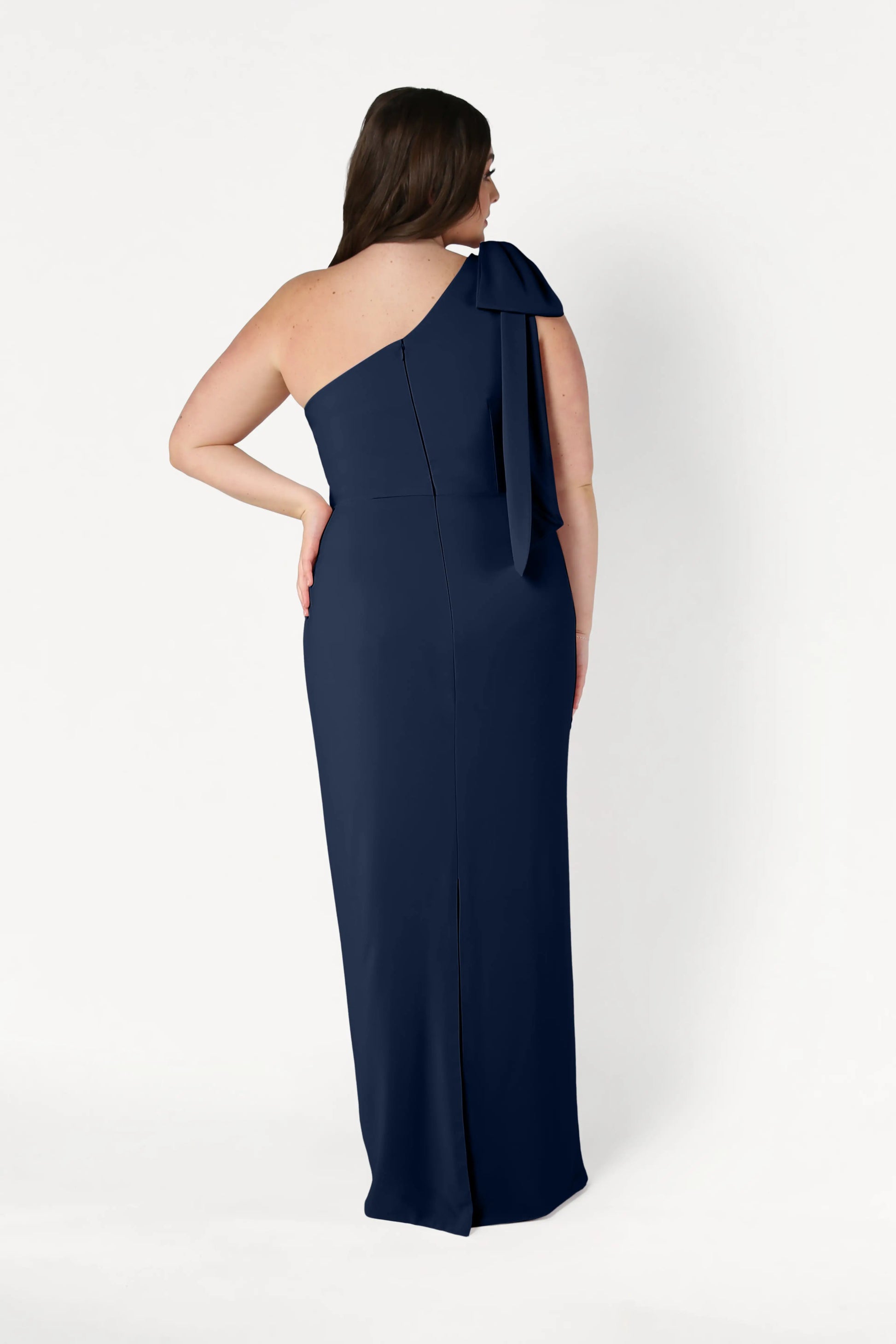 A woman faces away, showcasing the elegant back detail of the TH&TH Valentina Bridesmaid Dress in Navy, a floor-length crepe gown with a one-shoulder design and large bow, set against a plain white background.