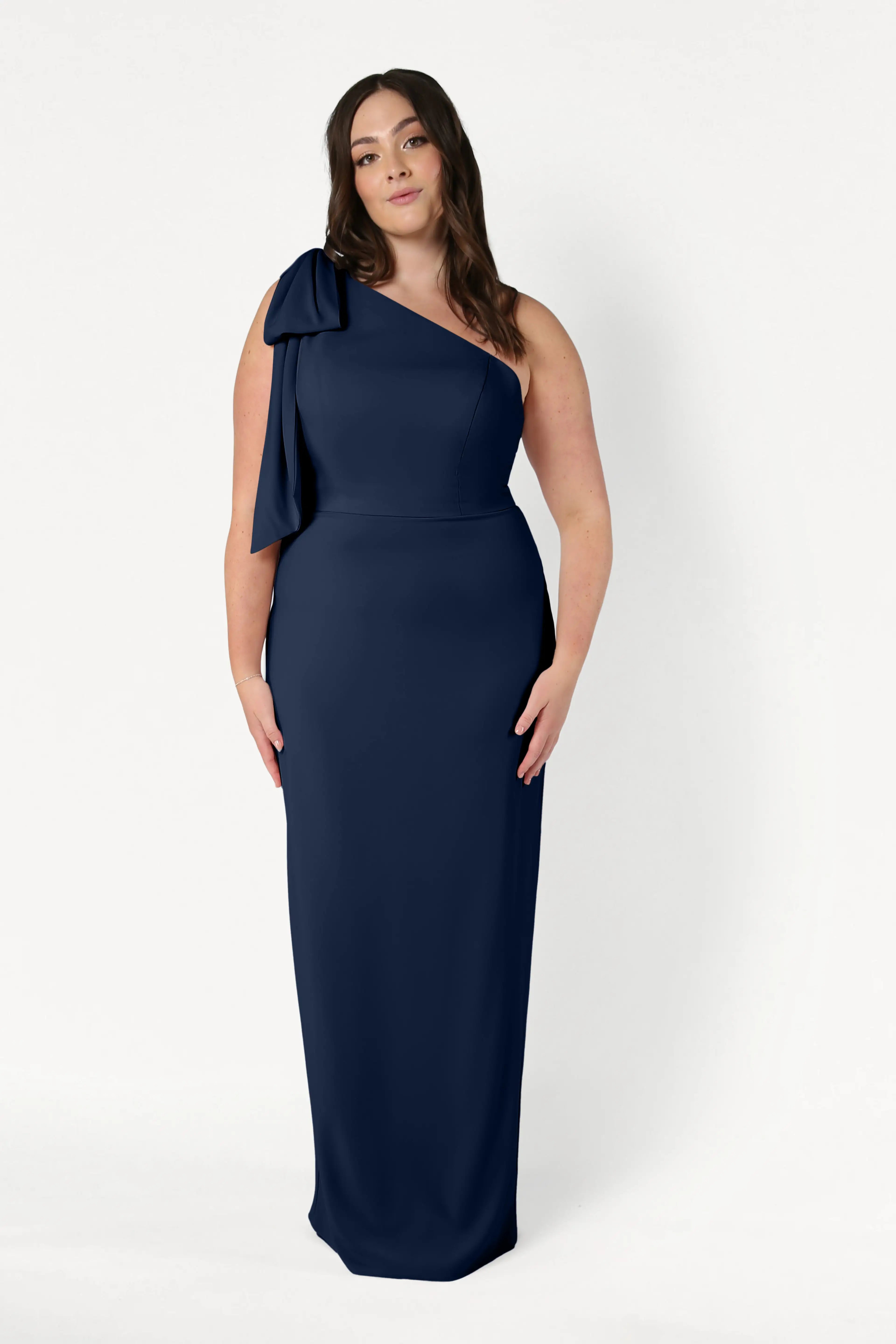 A woman with long brown hair and a neutral expression poses against a white background wearing the TH&TH Valentina Bridesmaid Dress in Navy, a one-shoulder crepe style featuring a bow on the shoulder.