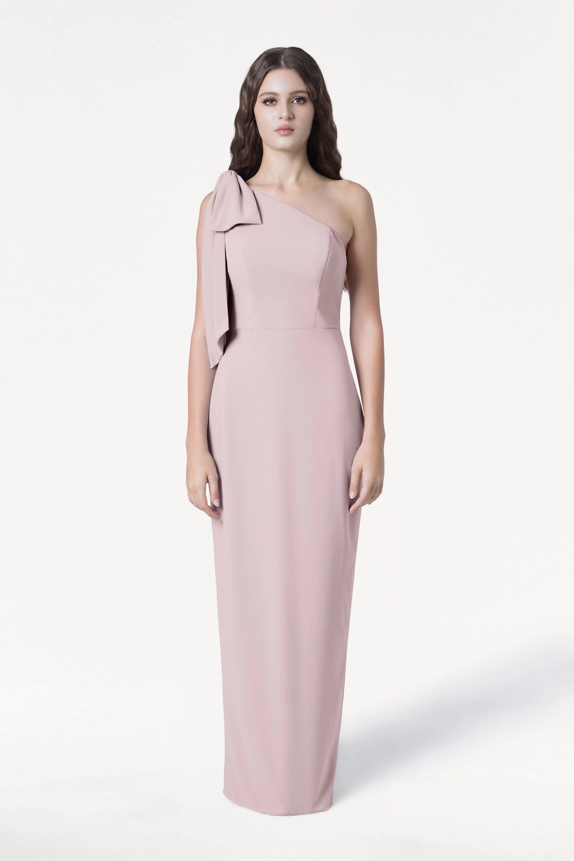 A woman faces forward in the TH&TH Valentina Bridesmaid Dress in Smoked Blush, featuring a modern crepe fabric and one-shoulder bow detail. She has long, wavy brown hair and a neutral expression against a plain white background.