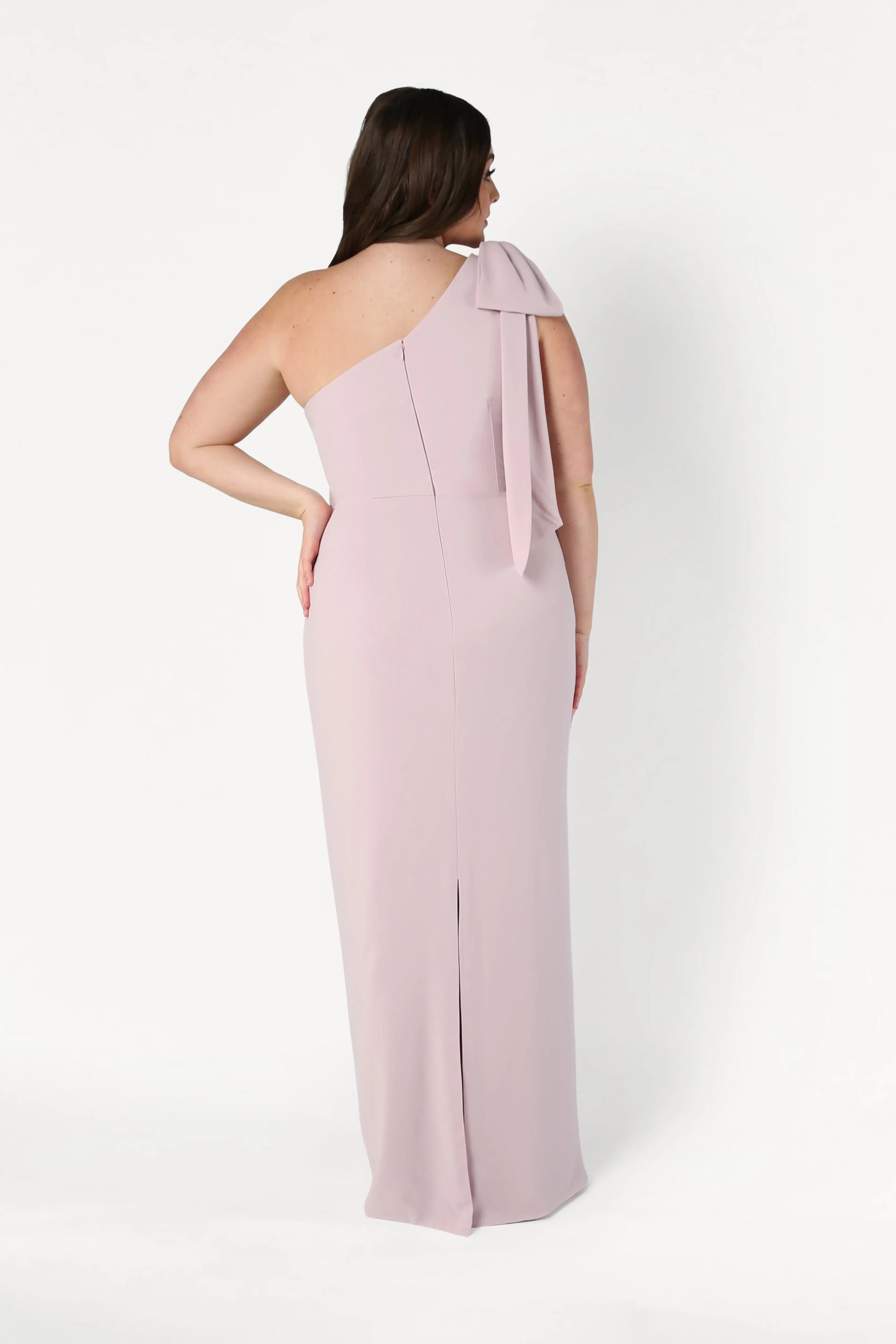 A woman faces away, wearing the TH&TH Valentina Bridesmaid Dress in Smoked Blush—a modern crepe gown with a draped one-shoulder detail and fitted silhouette, shown against a plain white background.