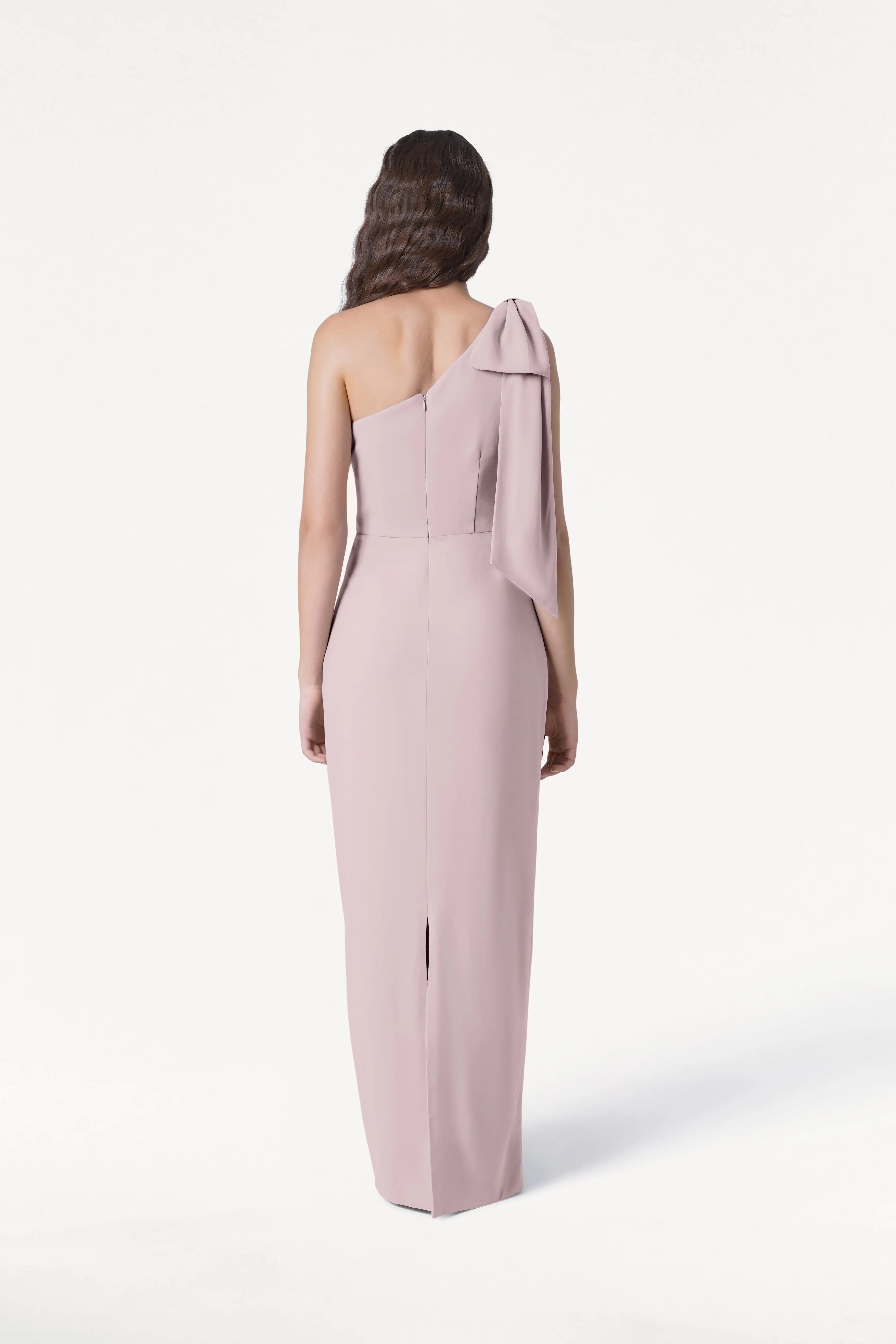 A woman with long, wavy brown hair stands facing away against a white background, wearing the TH&TH Valentina Bridesmaid Dress in Smoked Blush—featuring crepe fabric, a one-shoulder bow, and back slit.