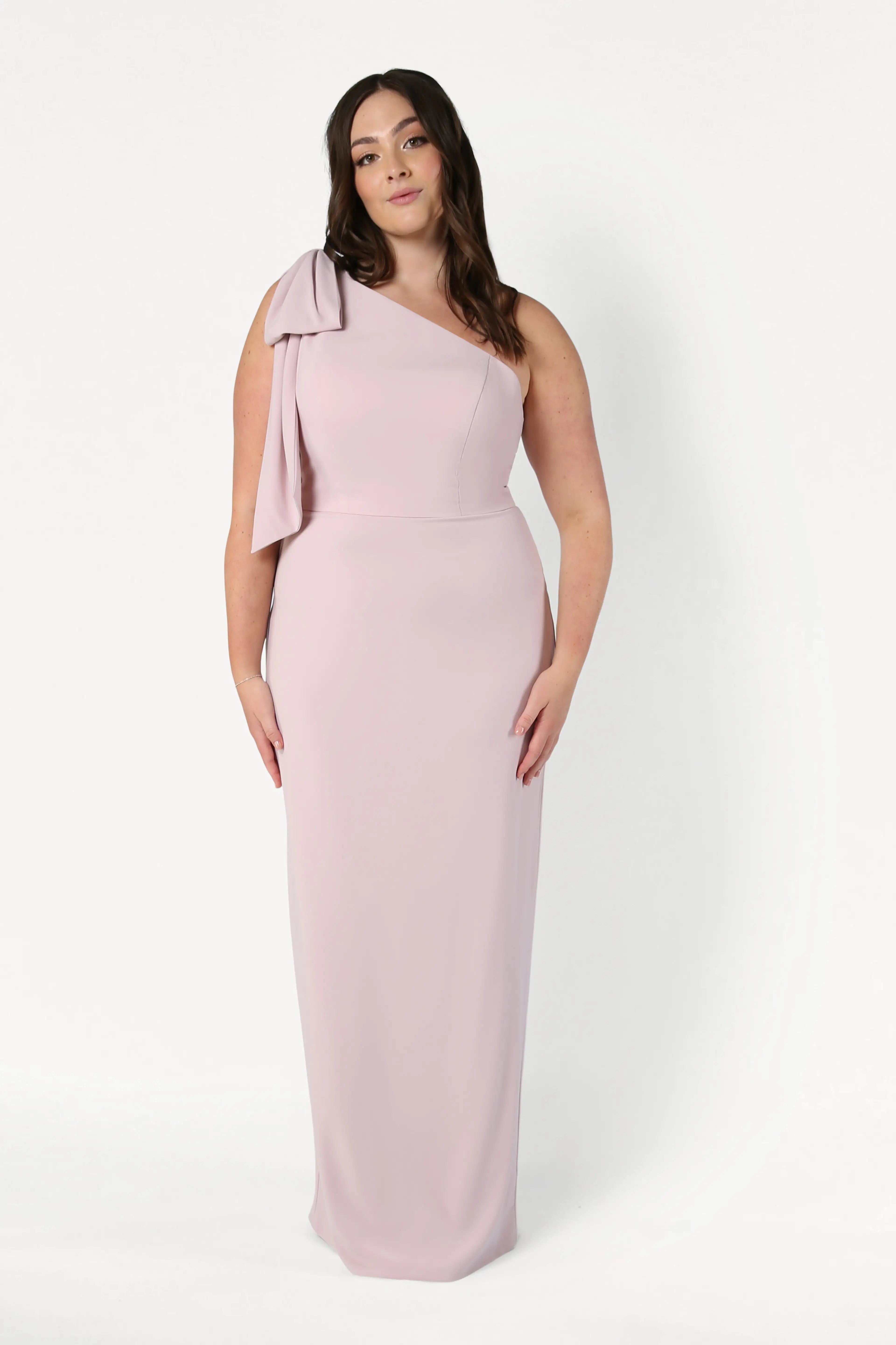 A woman wears the TH&TH Valentina Bridesmaid Dress in Smoked Blush, a floor-length, pale pink one-shoulder crepe gown with a shoulder bow, standing against a plain white background.