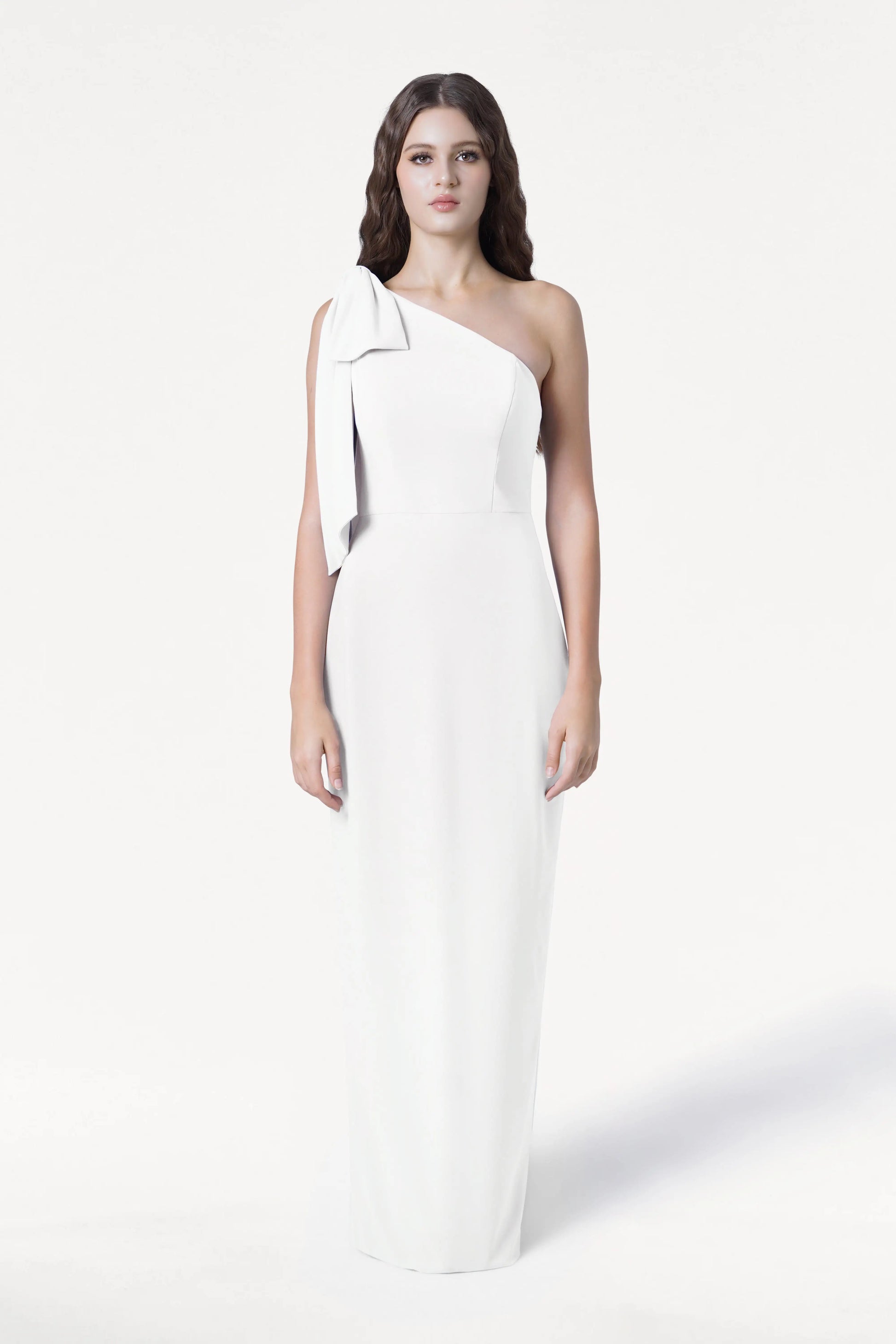 A woman with loose waves and a neutral expression models the TH&TH Valentina Dress in Ivory, a modern, size-inclusive crepe one-shoulder bridesmaid dress featuring a bow detail, against a white background.