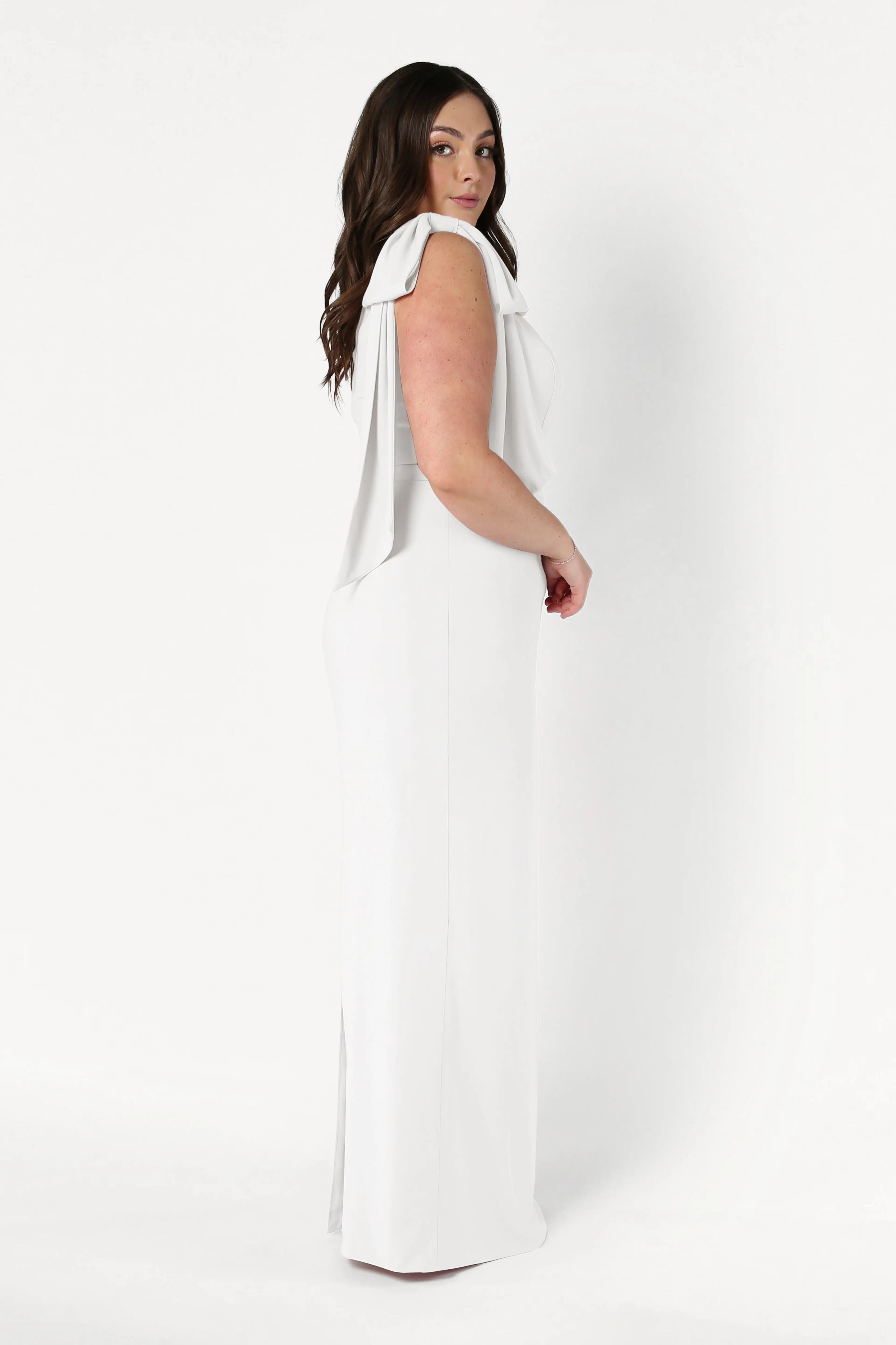 A woman with long brown hair stands sideways in the Valentina Dress in Ivory by TH&TH, featuring modern crepe fabric and draped one-shoulder details, posing gracefully against a plain white background.