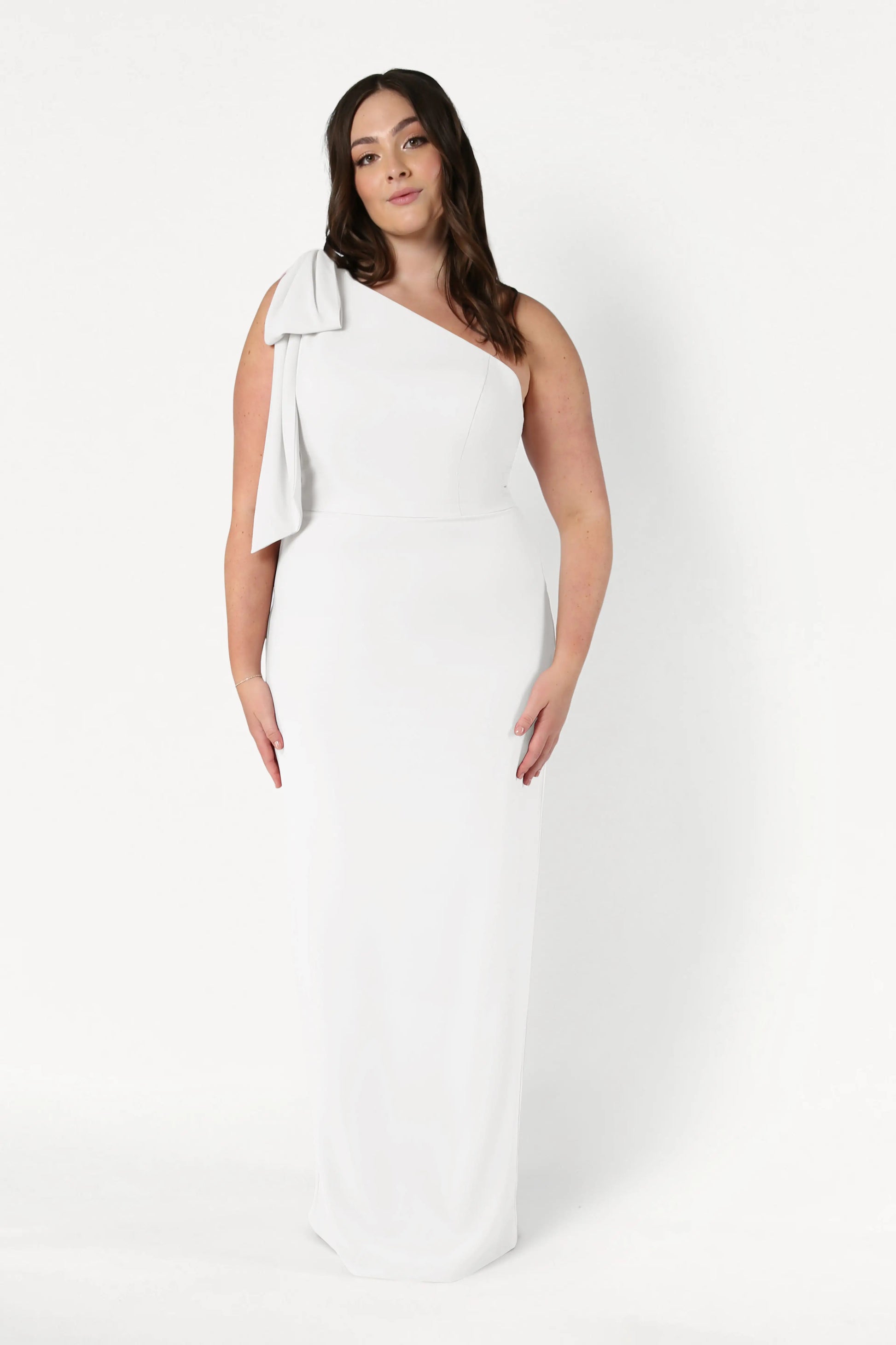 A woman stands against a white background wearing the Valentina Dress in Ivory by TH&TH, a long, elegant one-shoulder crepe bridesmaid dress with a bow detail. Her long, wavy dark hair frames her neutral expression.