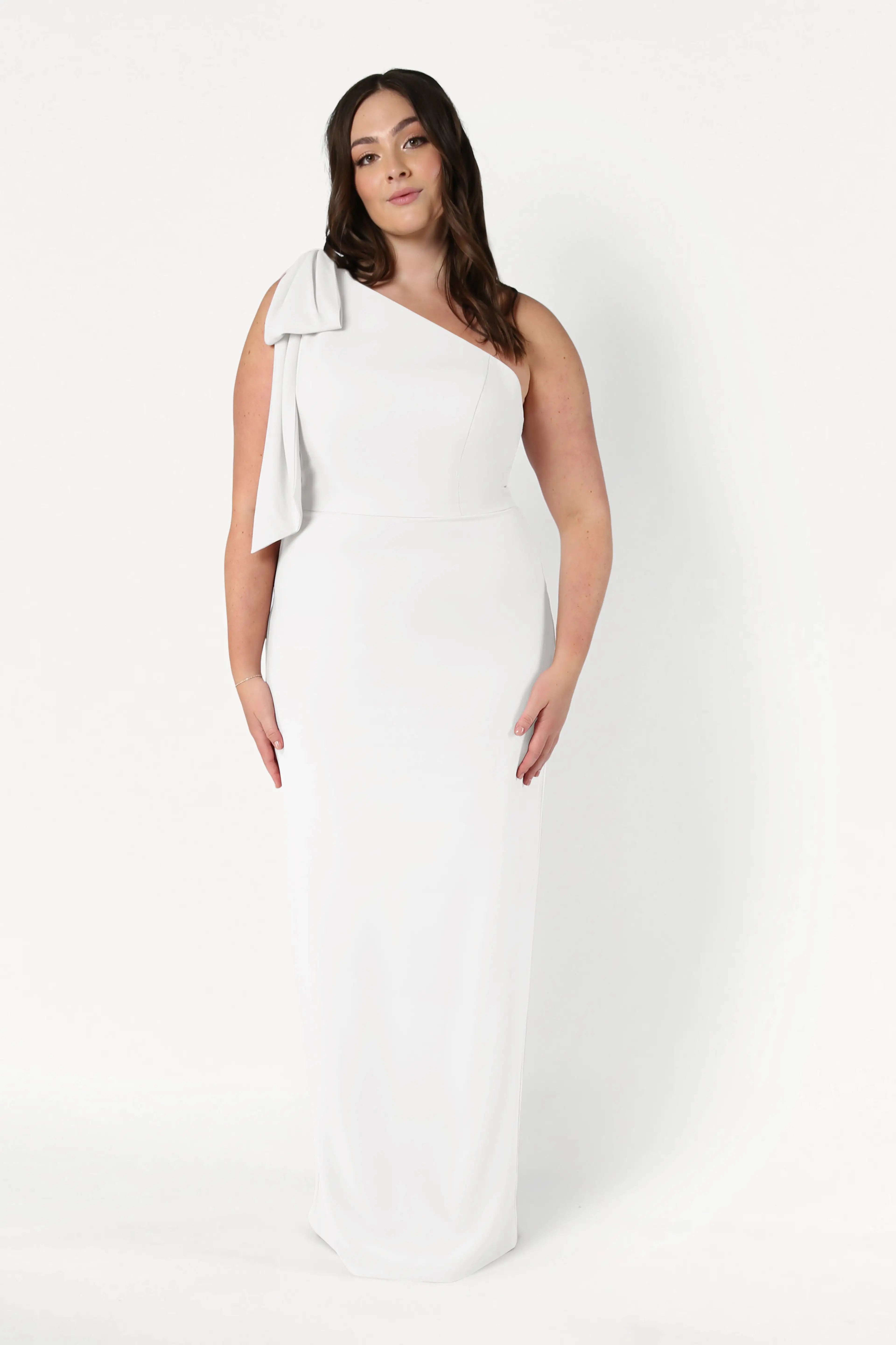 A woman stands against a white background wearing the Valentina Dress in Ivory by TH&TH, a long, elegant one-shoulder crepe bridesmaid dress with a bow detail. Her long, wavy dark hair frames her neutral expression.