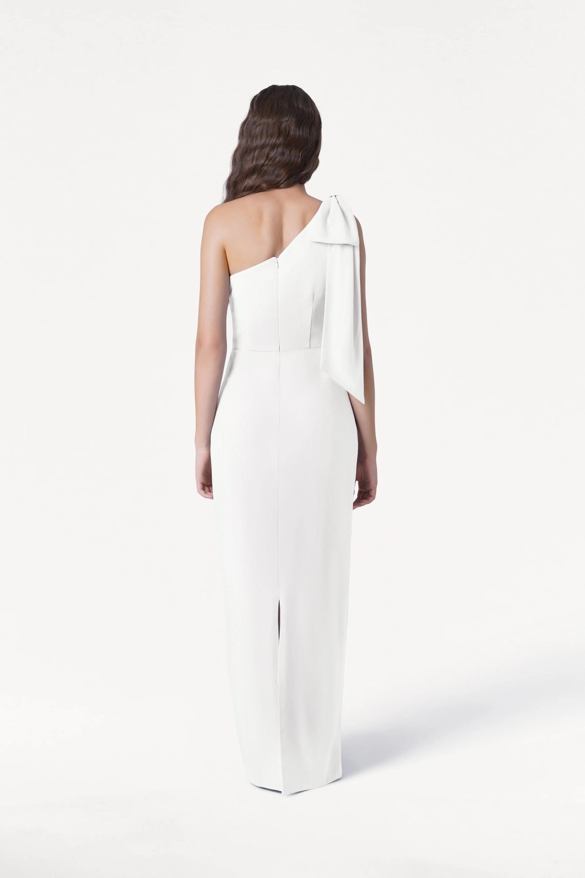 A woman with long wavy hair faces away, wearing the size-inclusive Valentina Dress in Ivory by TH&TH—a floor-length, one-shoulder bridesmaid dress with a bow detail—set against a plain white background.