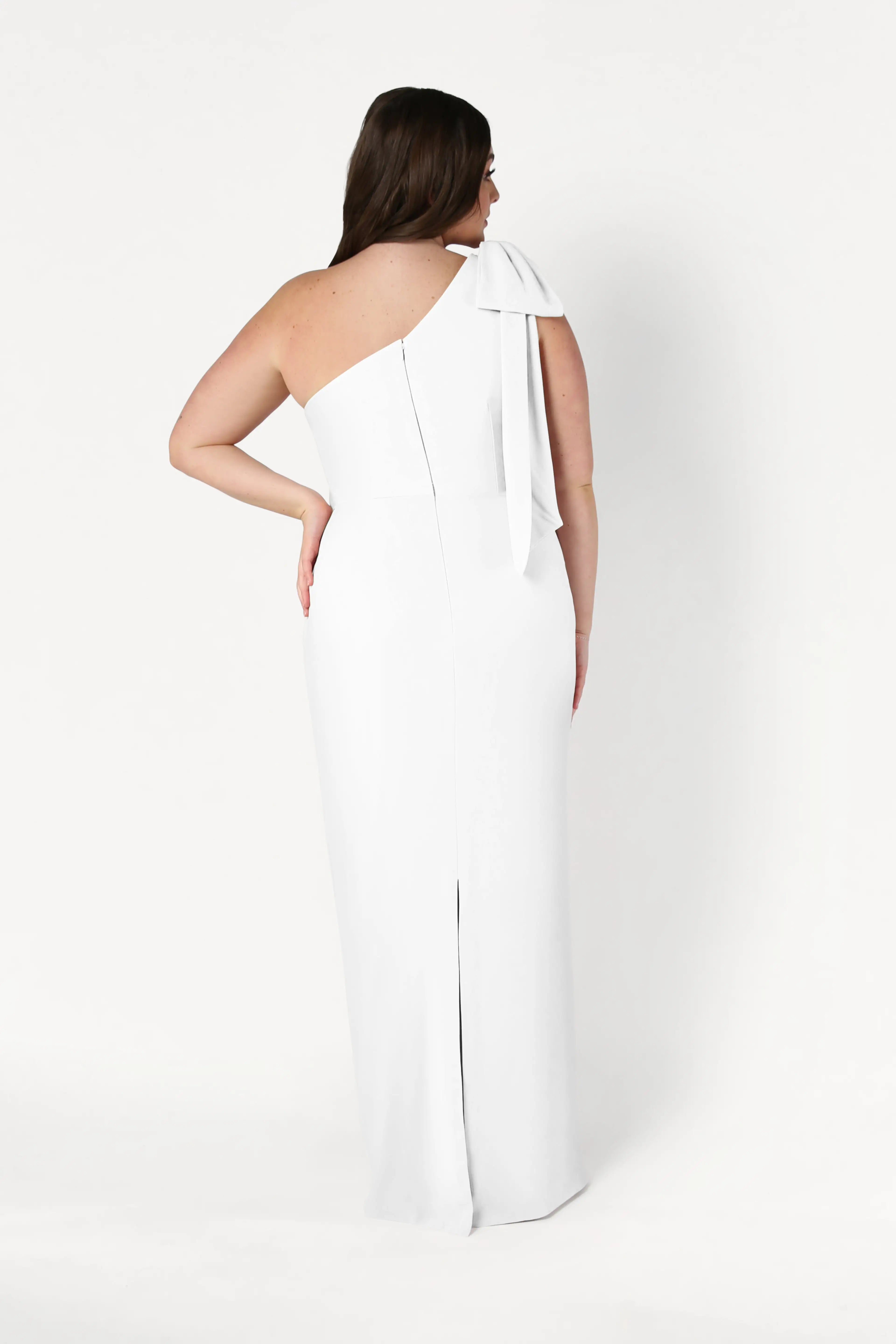 A woman stands facing away, modeling the TH&TH Valentina Dress in Ivory, a size-inclusive, long one-shoulder crepe gown with a shoulder bow and back slit. She poses against a plain white background.