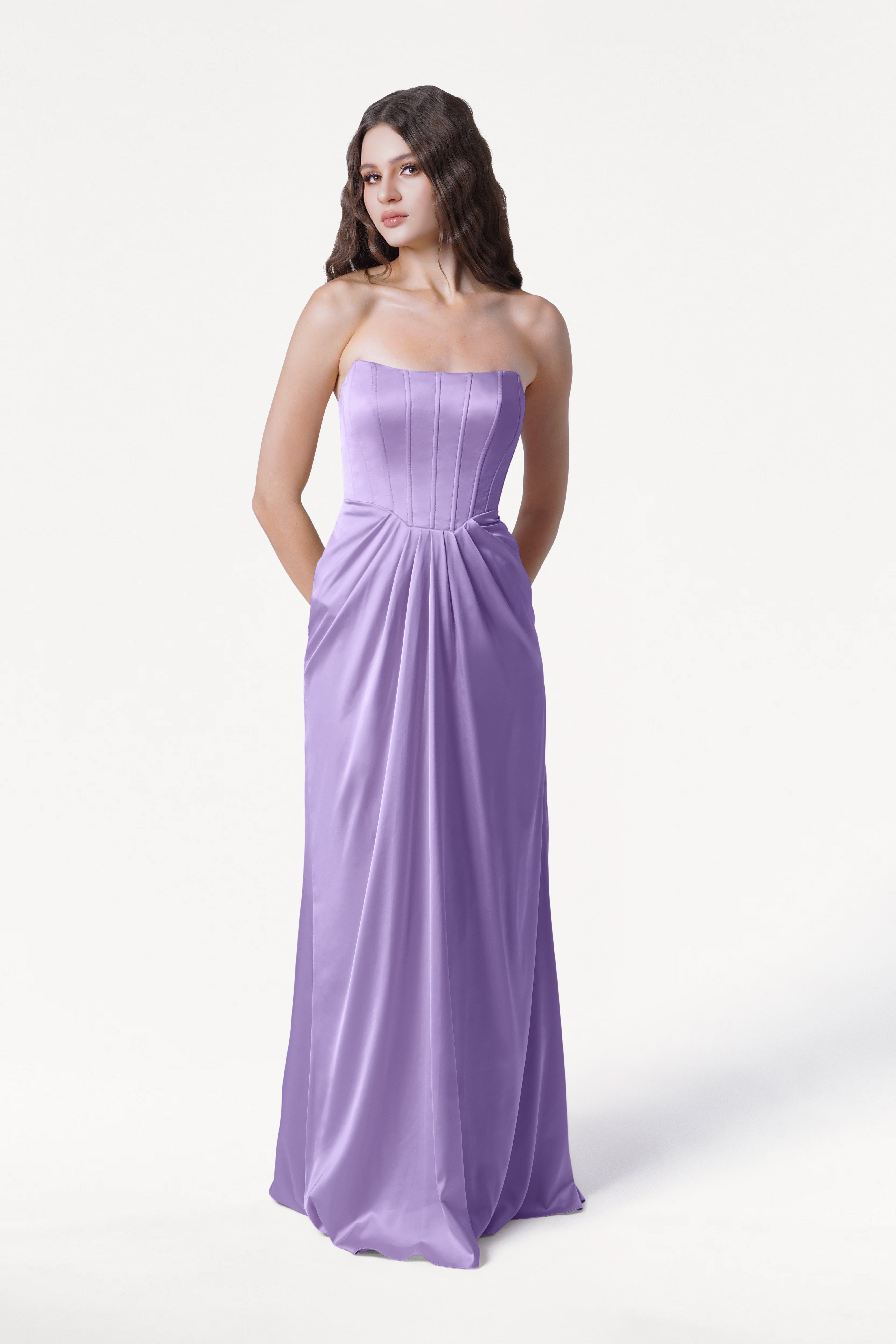 A woman with long brown hair models the TH&TH Selena Bridesmaid Dress in Violet Satin, a strapless, floor-length gown with a fitted bodice and subtle waist pleating, standing against a plain white background.