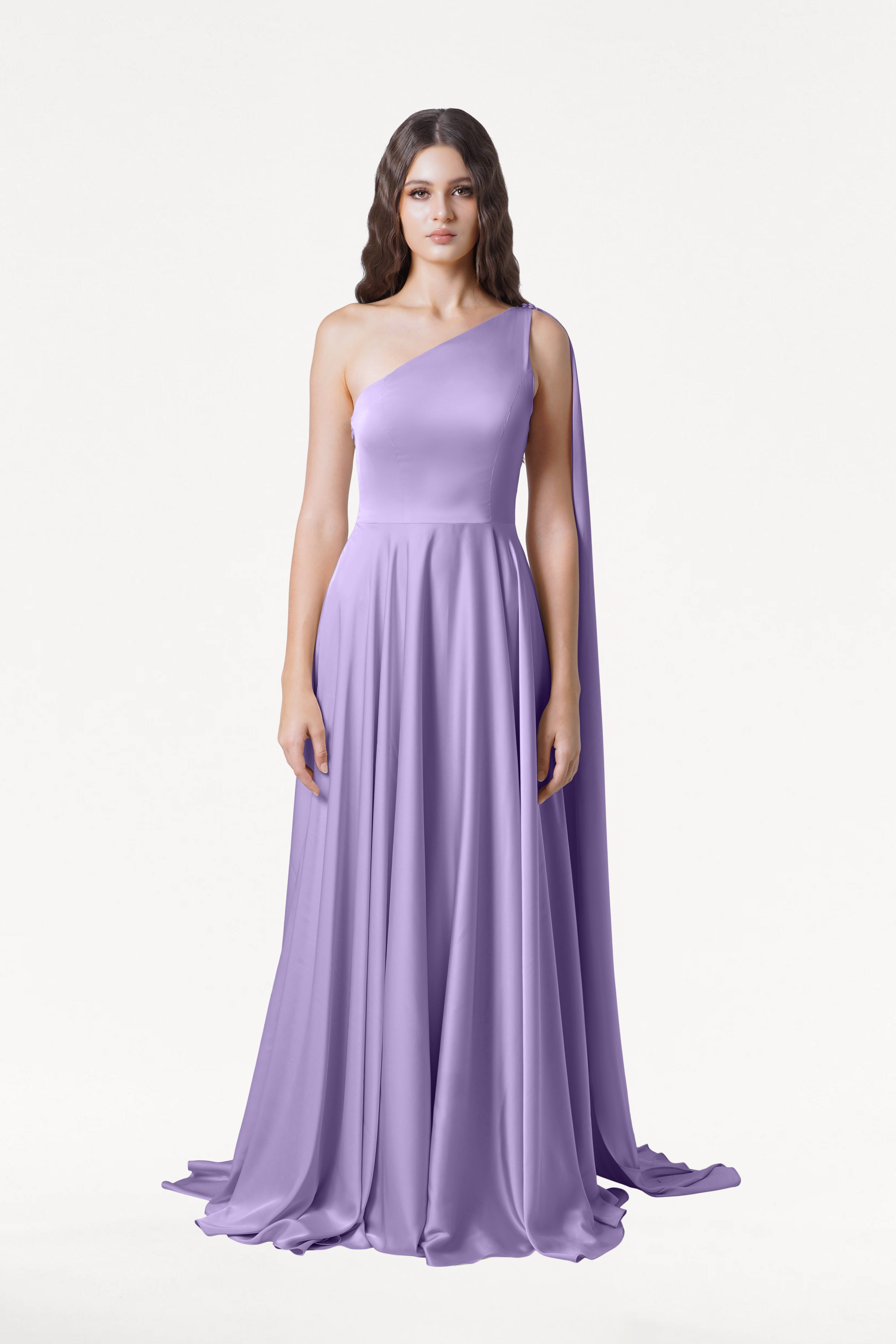 A woman with long wavy brown hair wears the Lyra Bridesmaid Dress in Violet Satin by TH&TH, featuring a one-shoulder strap and draped cape detail. She faces forward with a neutral expression against a plain white background.