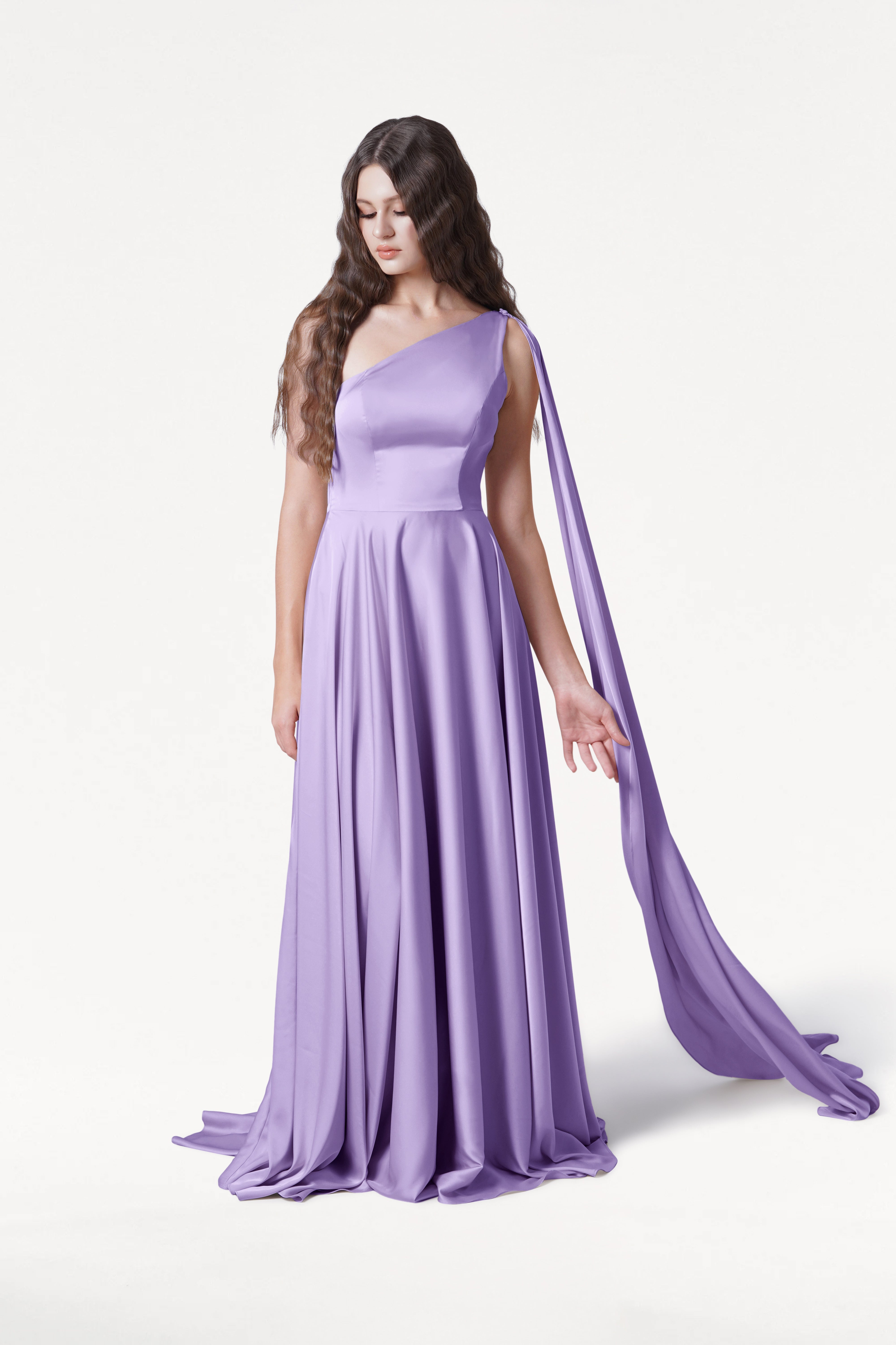 A woman with long, wavy brown hair wears the TH&TH Lyra Bridesmaid Dress in Violet Satin—a size-inclusive, floor-length one-shoulder gown with a flowing sash—while standing against a plain white background, looking down with an arm gently extended.