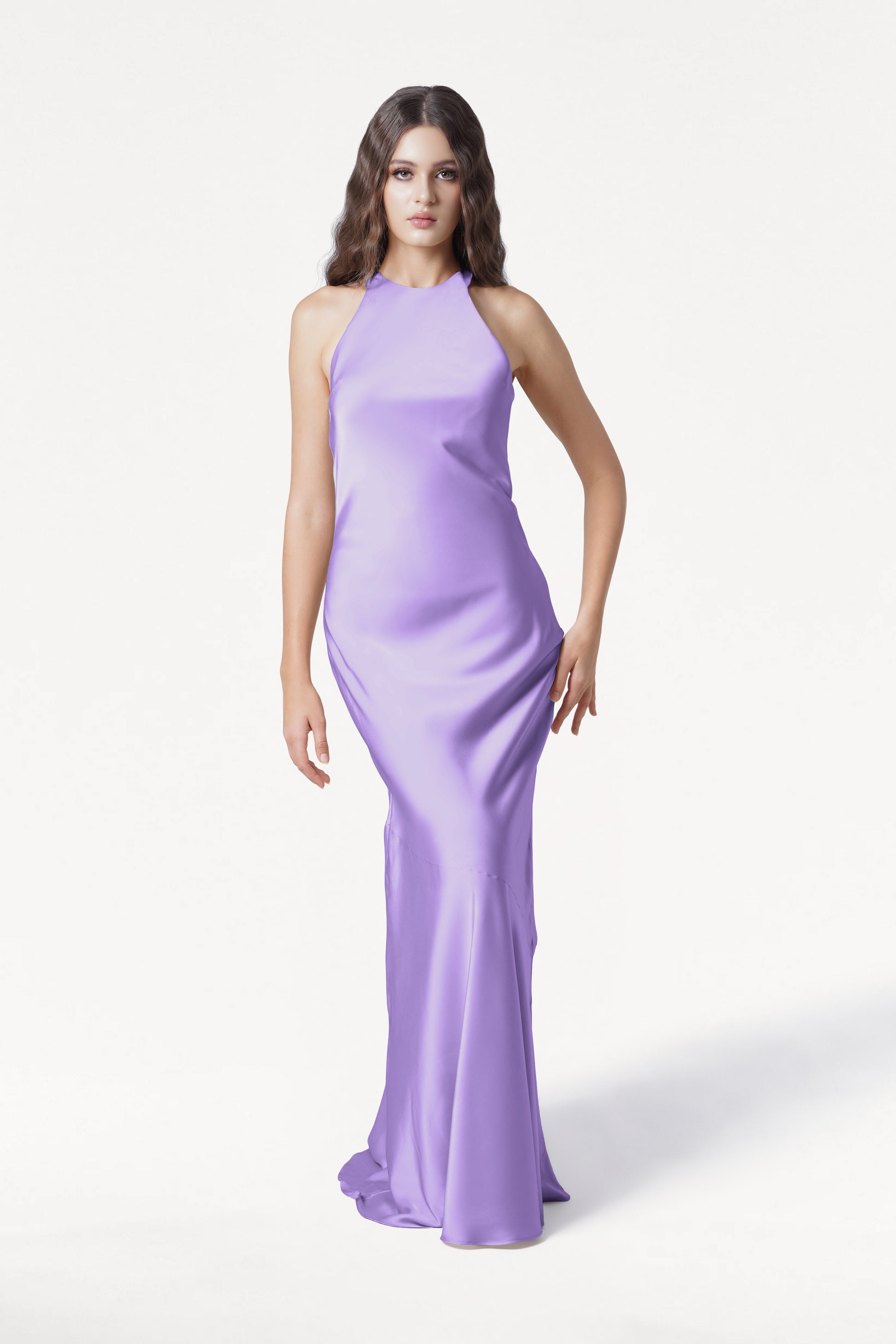 A woman with long wavy brown hair stands against a white background, wearing the TH&TH Mira Bridesmaid Dress in violet satin—a sleeveless, floor-length gown with a high neckline.