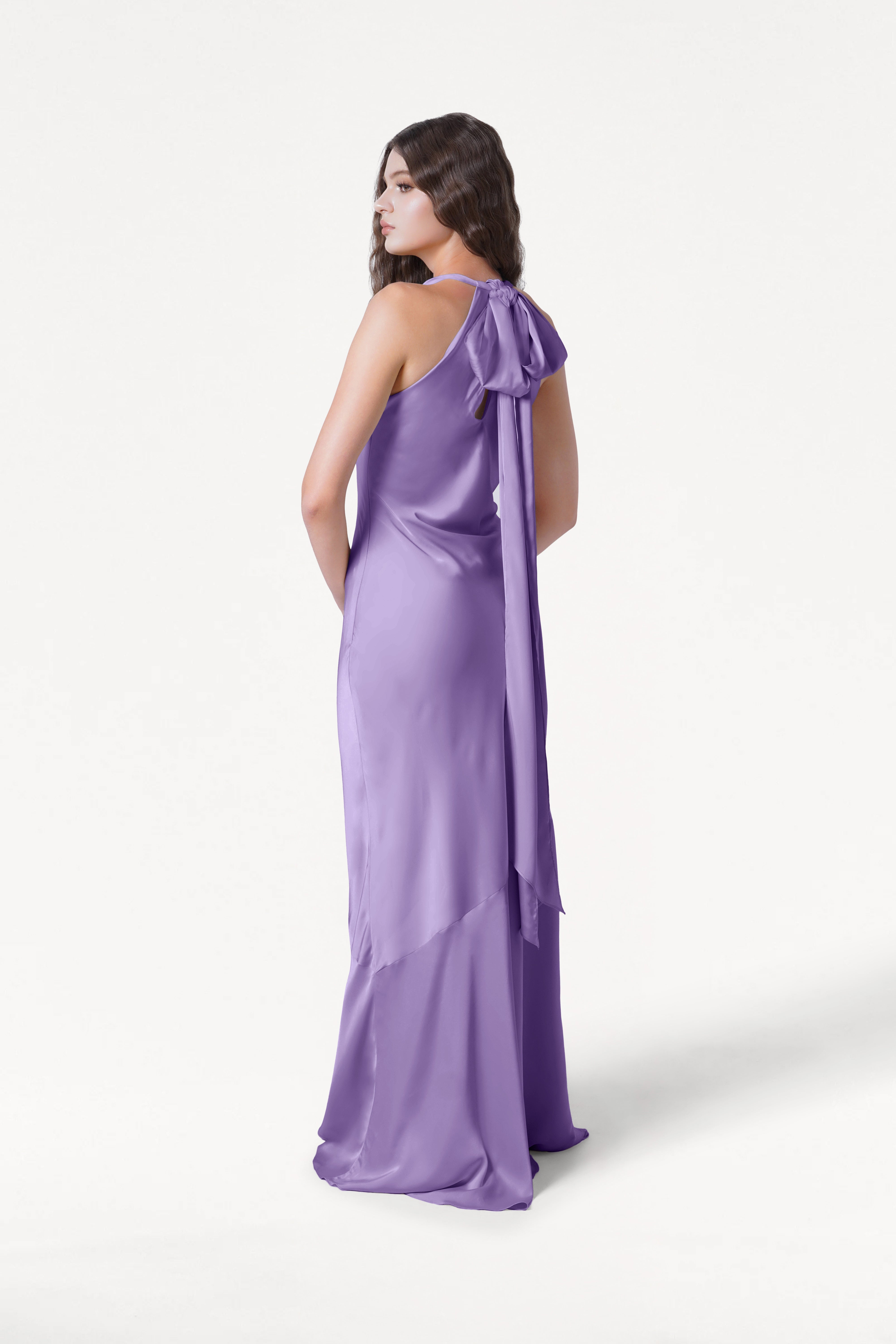 A woman with wavy brown hair stands against a white background wearing the TH&TH Mira Bridesmaid Dress in violet satin, a floor-length halterneck style with a long bow at the back of the neck.