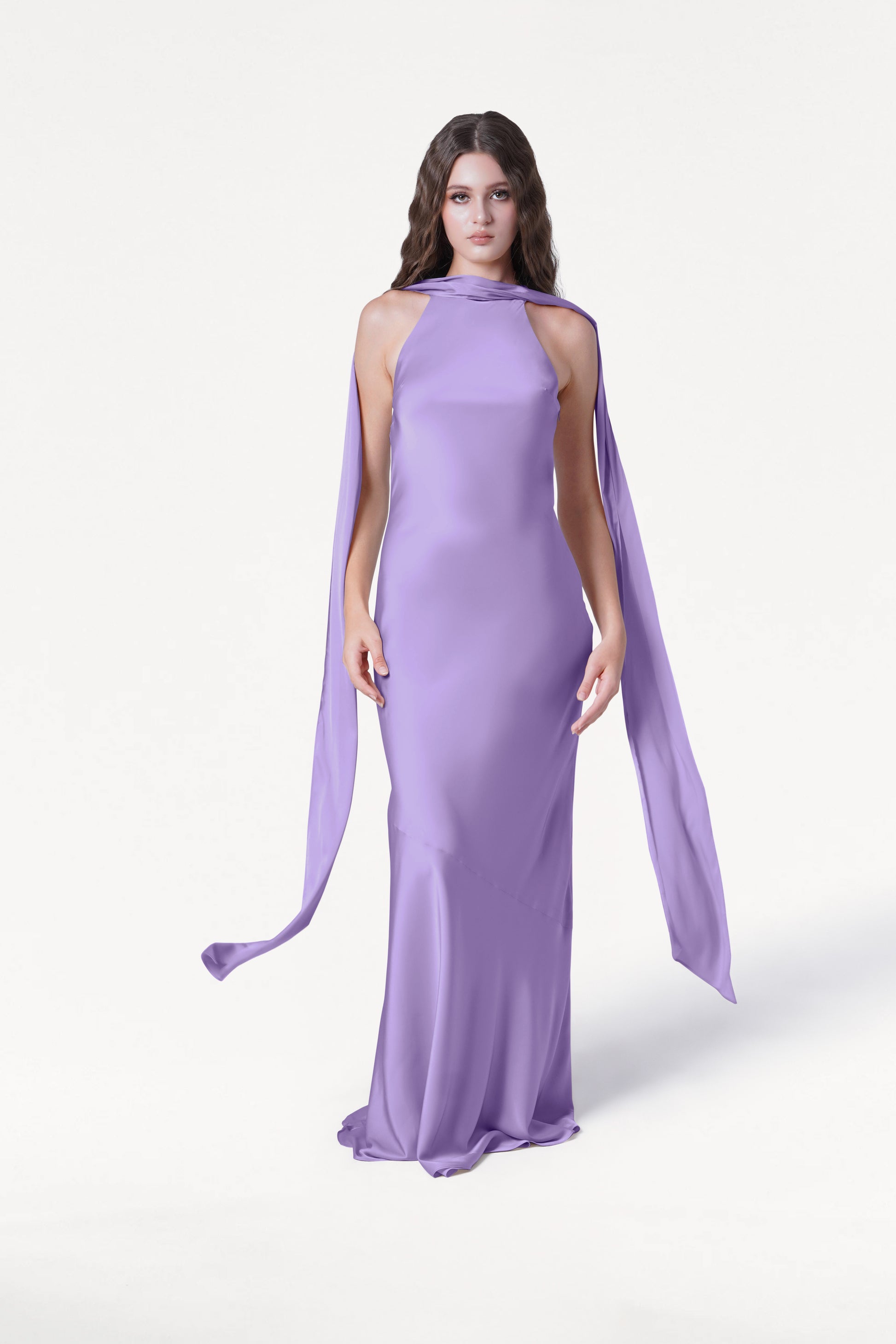 A woman with long brown hair models the TH&TH Mira Bridesmaid Dress in Violet Satin, a sleeveless, floor-length halterneck gown, standing against a plain white background.