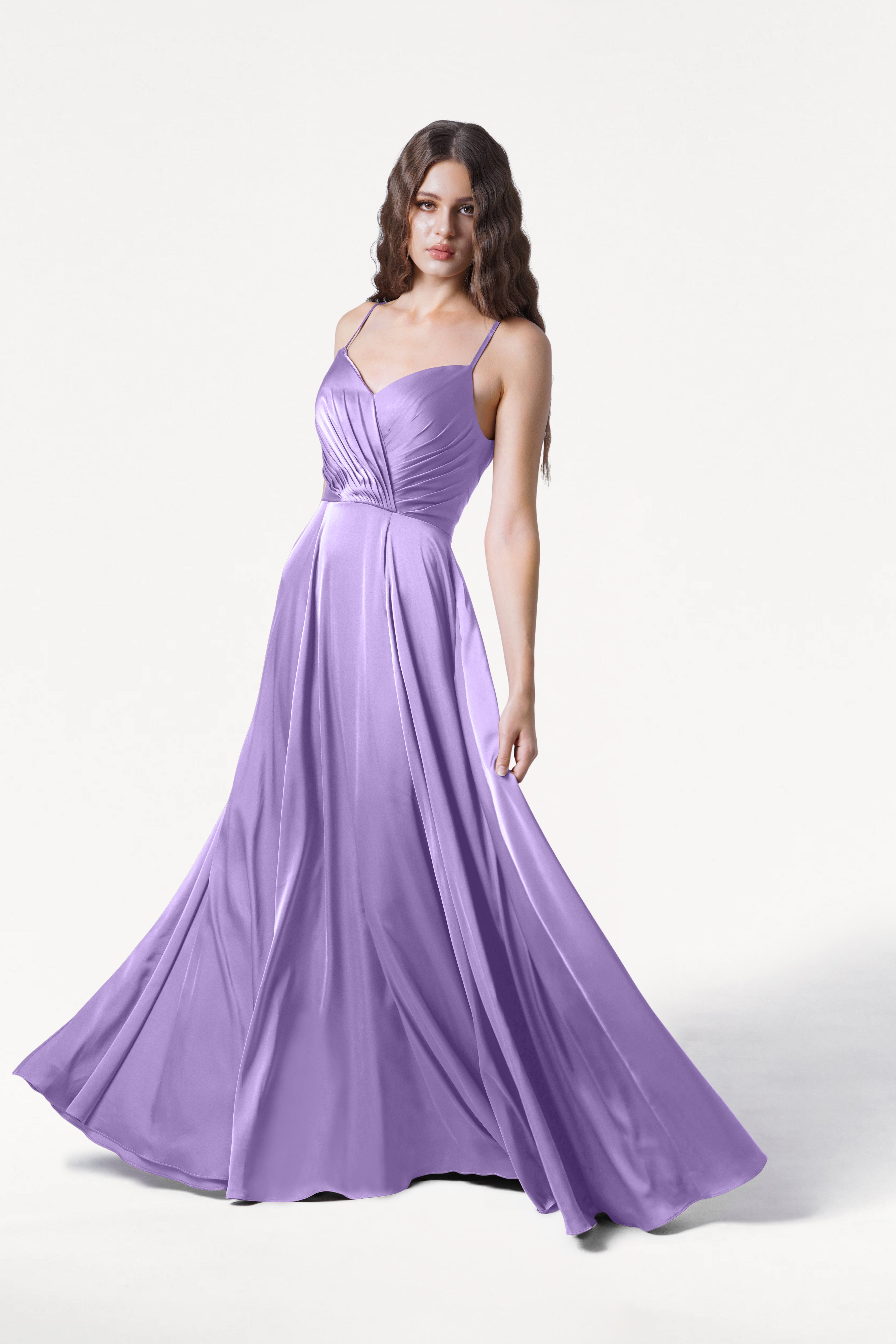 A woman stands against a plain white background wearing the TH&TH Isla Bridesmaid Dress in Violet Satin—a long, flowing dress with thin straps and a pleated bodice.
