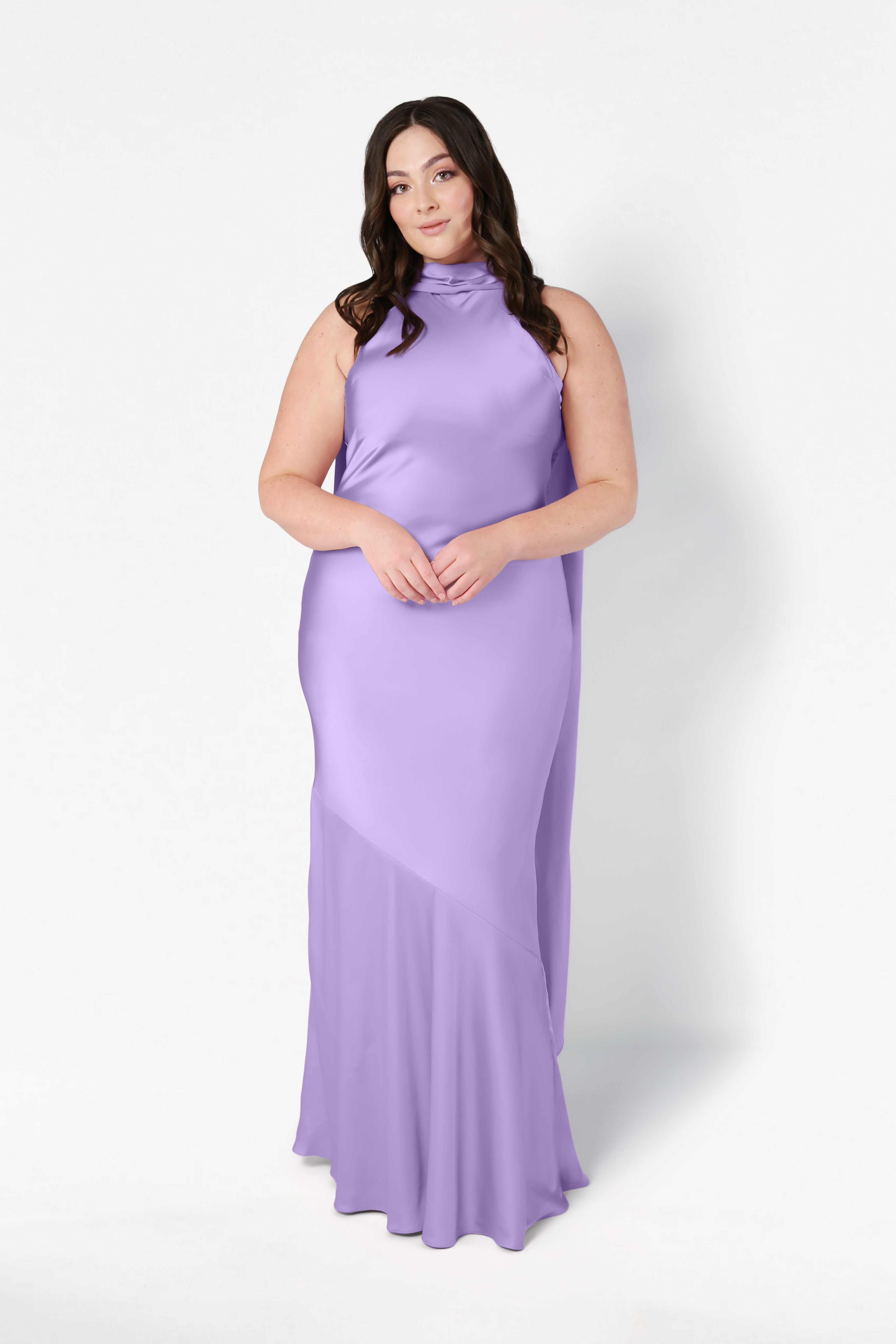 A woman with long dark hair stands against a white background wearing the TH&TH Mira Bridesmaid Dress in floor-length violet satin, sleeveless with a high neckline. She faces forward, hands gently clasped in front of her.