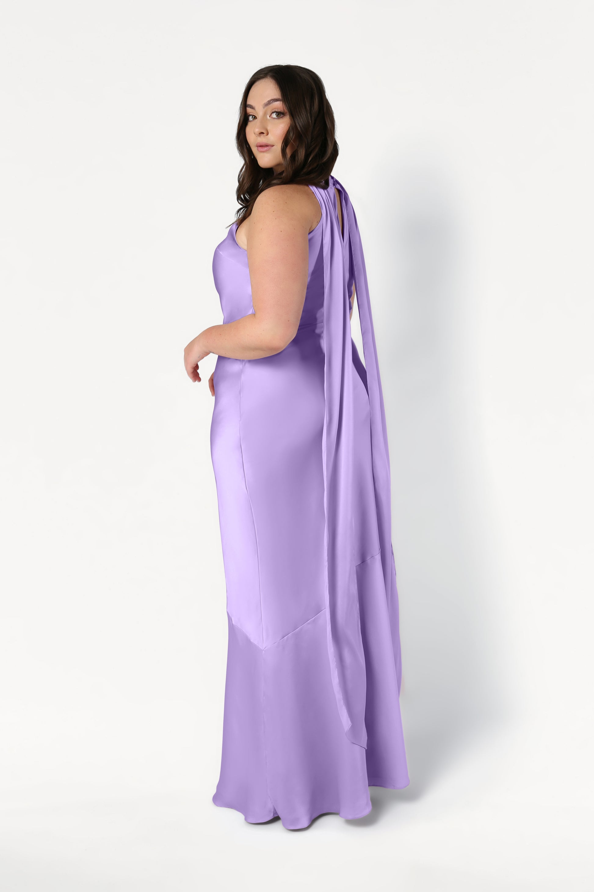 A woman in the Mira Bridesmaid Dress in Violet Satin by TH&TH, featuring a long, sleeveless design and flowing cape, stands against a white background, looking over her shoulder at the camera.