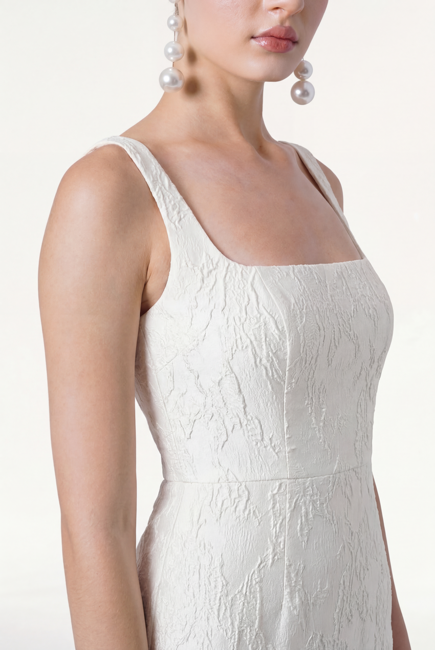 A woman wears the Waldorf Bridal Dress in Vintage Ivory Brocade by TH&TH—a textured white sleeveless gown with a square neckline—paired with large dangling pearl earrings, shown from shoulders to mid-torso against a plain light background.