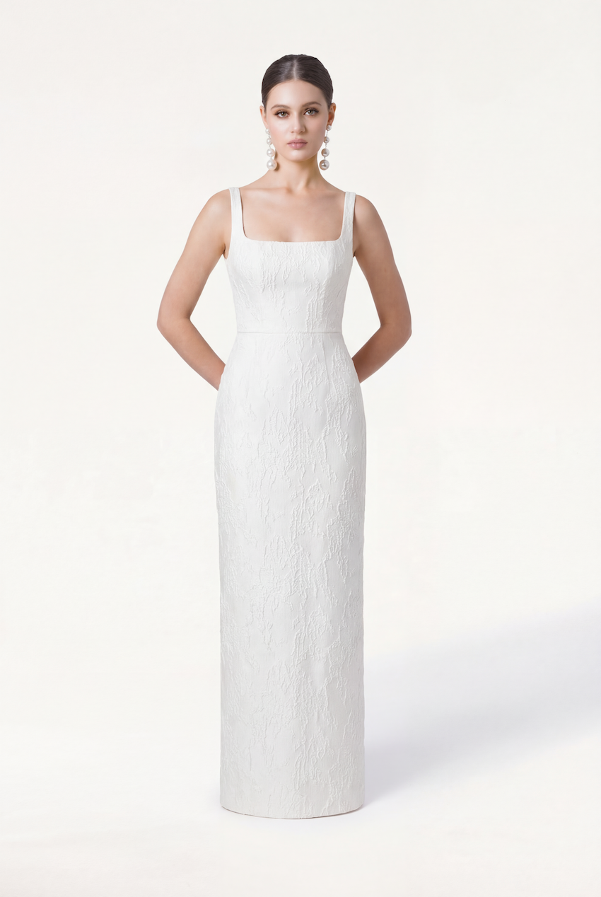 A woman stands against a plain light background, hands behind her back, wearing statement earrings and the TH&TH Waldorf Bridal Dress in Vintage Ivory Brocade—a sleeveless, floor-length gown with a square neckline and subtle texture.