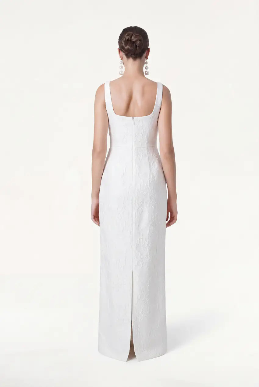 A woman with dark hair in a low bun models the TH&TH Waldorf Bridal Dress in Vintage Ivory Brocade, featuring a sleeveless, floor-length column silhouette, square neckline, back zipper and slit. She wears large drop earrings against a white background.