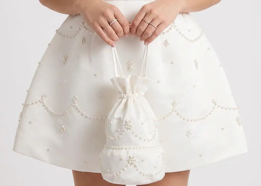 A person in a pearl-adorned white dress holds the TH&TH 'Wedding Cake' Embellished Bag in Ivory—a white drawstring bag with delicate pearls. The image shows only the torso and hands against a plain background.