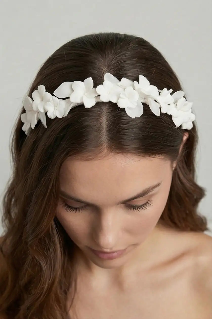 A woman with long brown hair wears the Windsor Ceramic Floral Headband by TH&TH, featuring delicate white flowers. She looks down, her loose waves framing her face against a plain background that highlights the elegant bridal accessory.