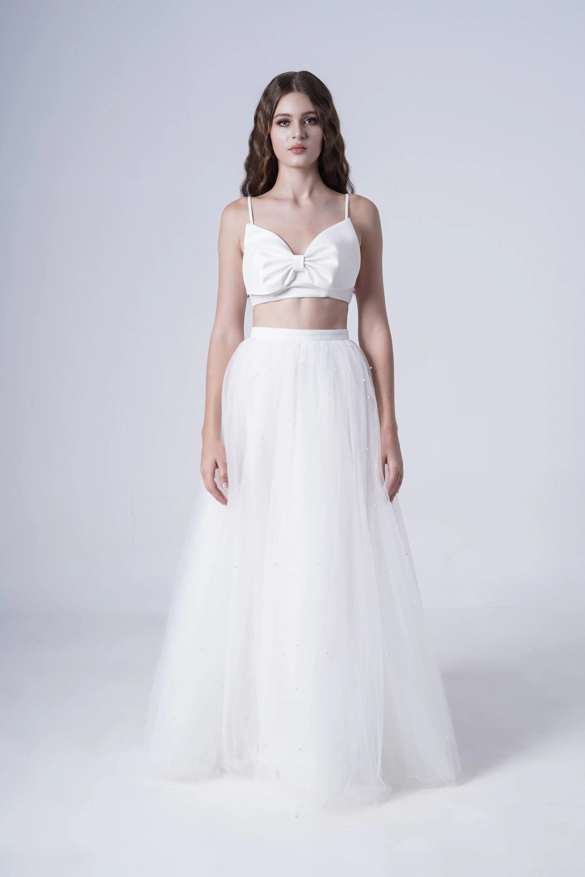 A woman poses against a white background in the TH&TH Wyatt Bridal Skirt in Ivory Tulle, styled with a knotted crop top. Her hair falls in loose waves and she maintains a neutral expression.