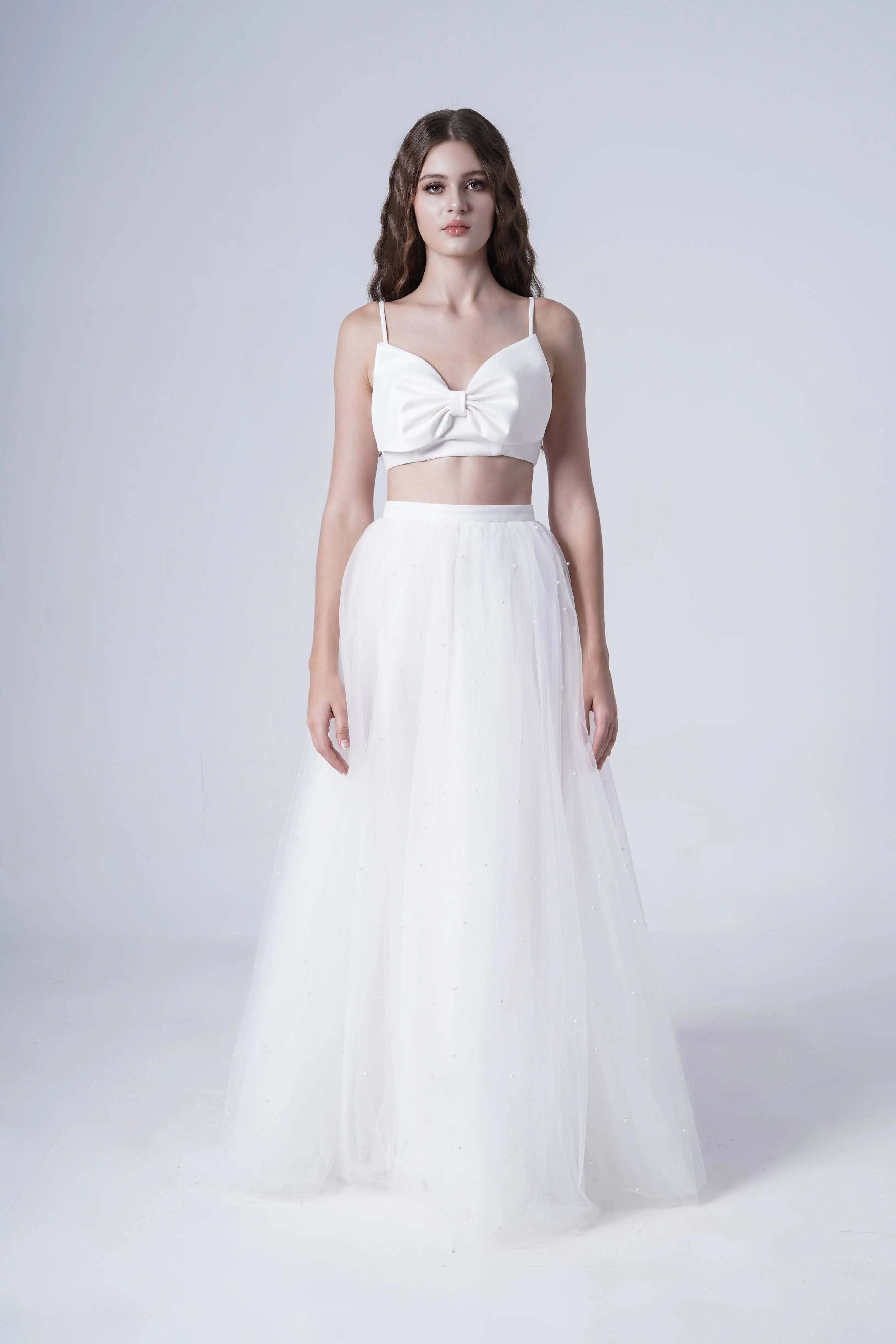 A woman poses against a white background in the TH&TH Wyatt Bridal Skirt in Ivory Tulle, styled with a knotted crop top. Her hair falls in loose waves and she maintains a neutral expression.