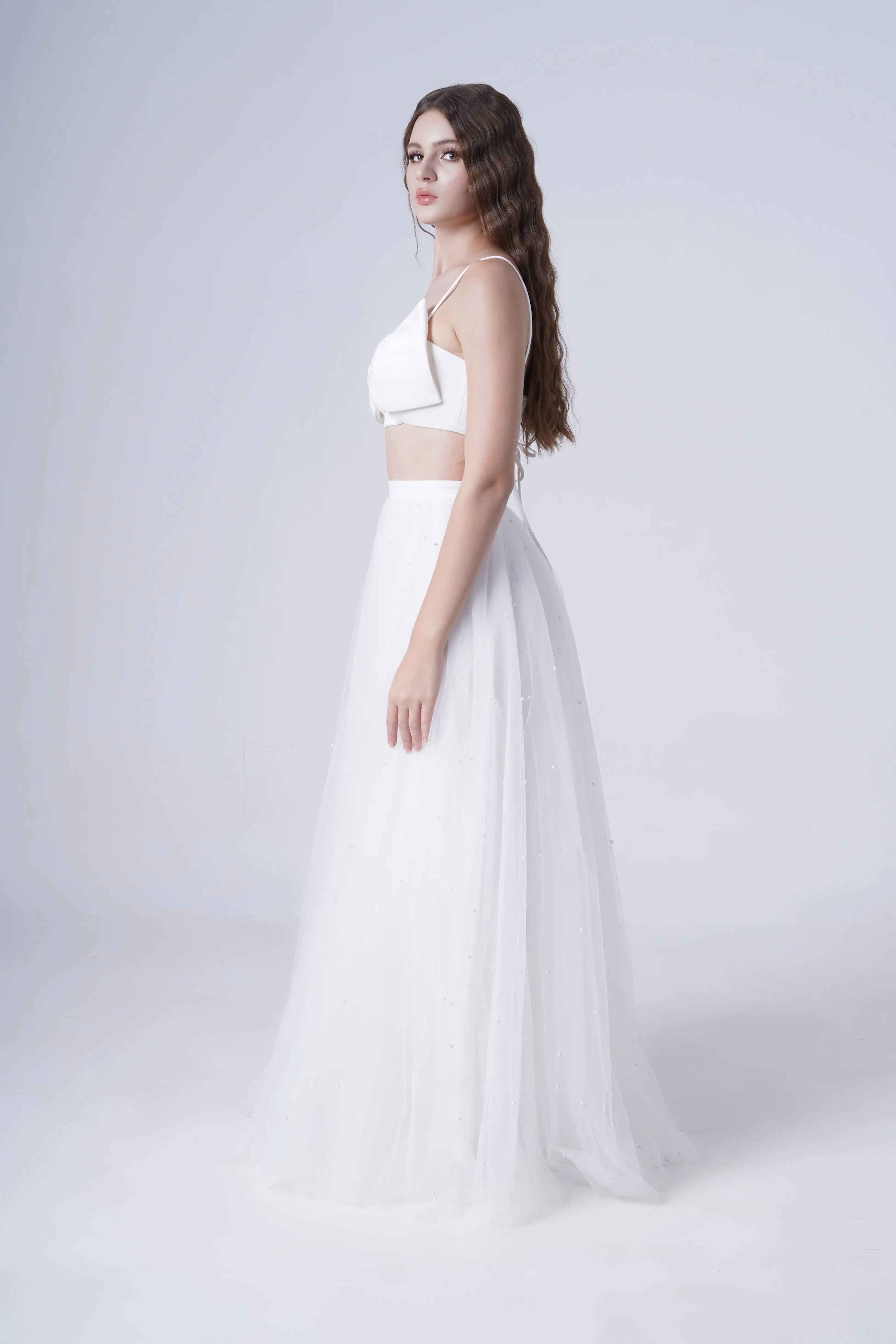A young woman with long wavy hair stands in profile, wearing the TH&TH Wyatt Bridal Skirt in Ivory Tulle and a white crop top, exuding the modern elegance of bridal separates against a plain light background.