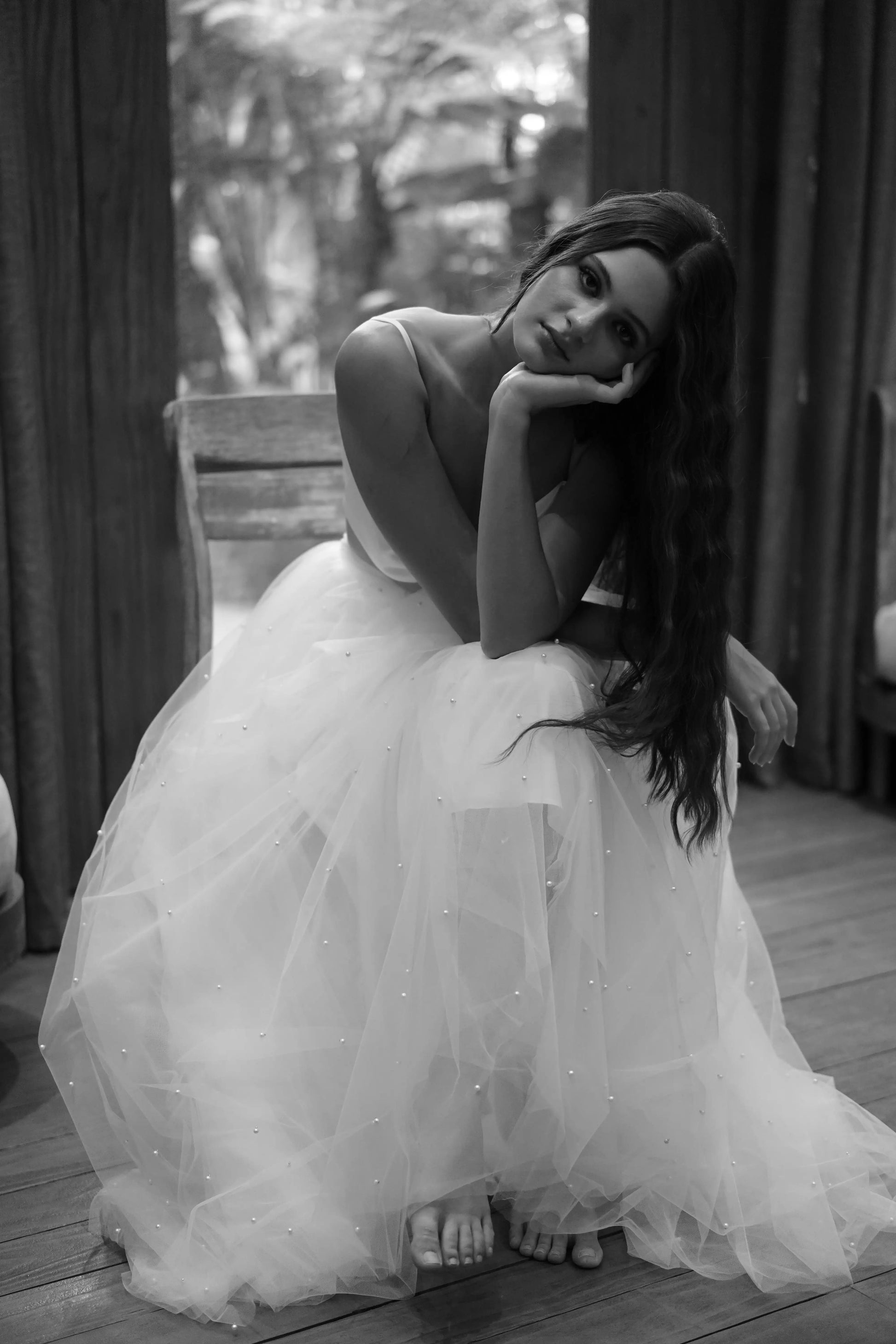 A woman wearing the TH&TH Wyatt Bridal Skirt in Ivory Tulle sits barefoot on a wooden chair indoors, resting her head on her hand and looking at the camera in this black and white photo.