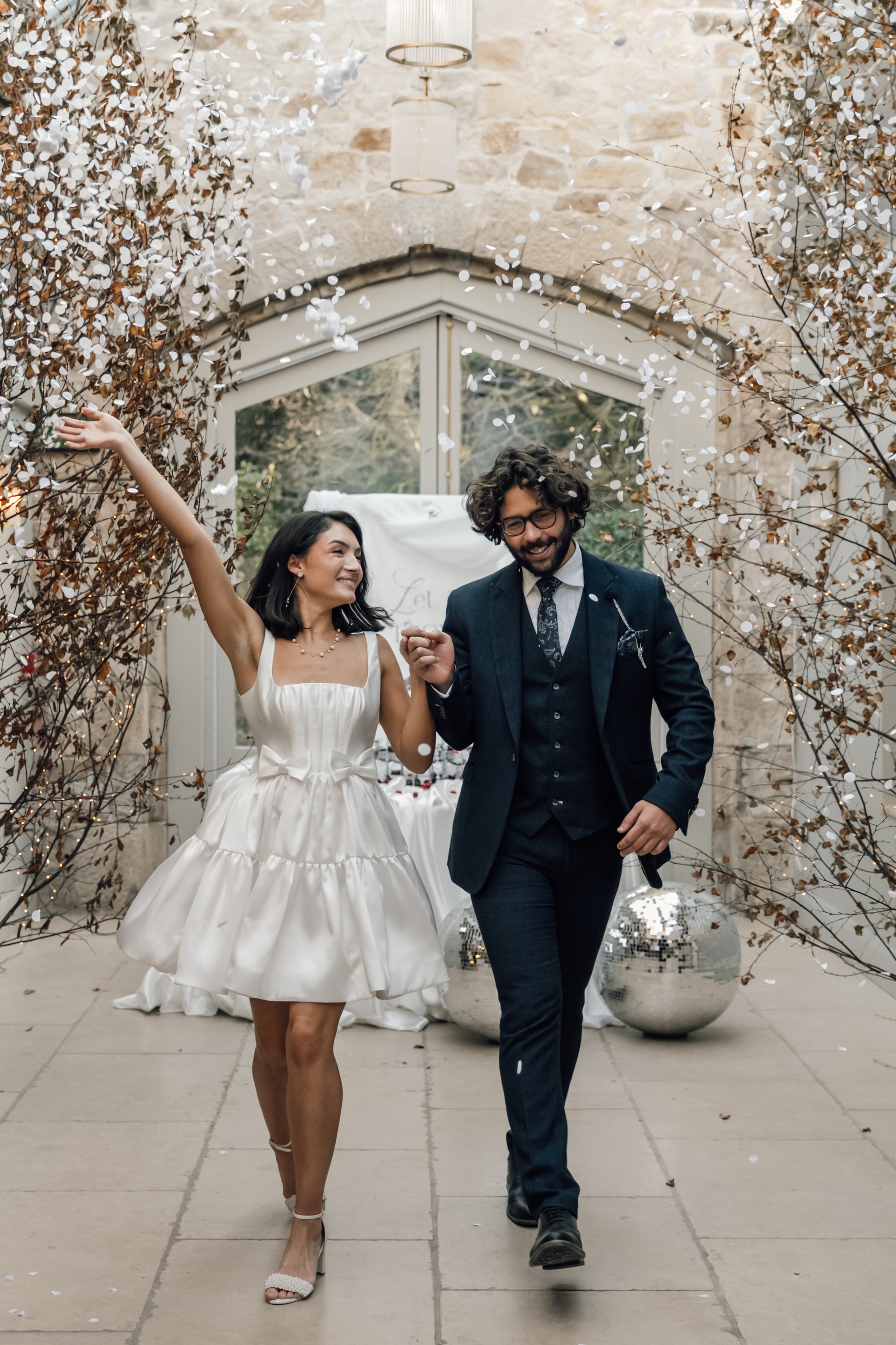 A joyful couple walks hand in hand down an aisle with white confetti. The woman wears the TH&TH Bloom Bridal Mini Dress in Ivory, featuring a corseted bodice, and the man is dressed in a dark suit against a stone wall backdrop.