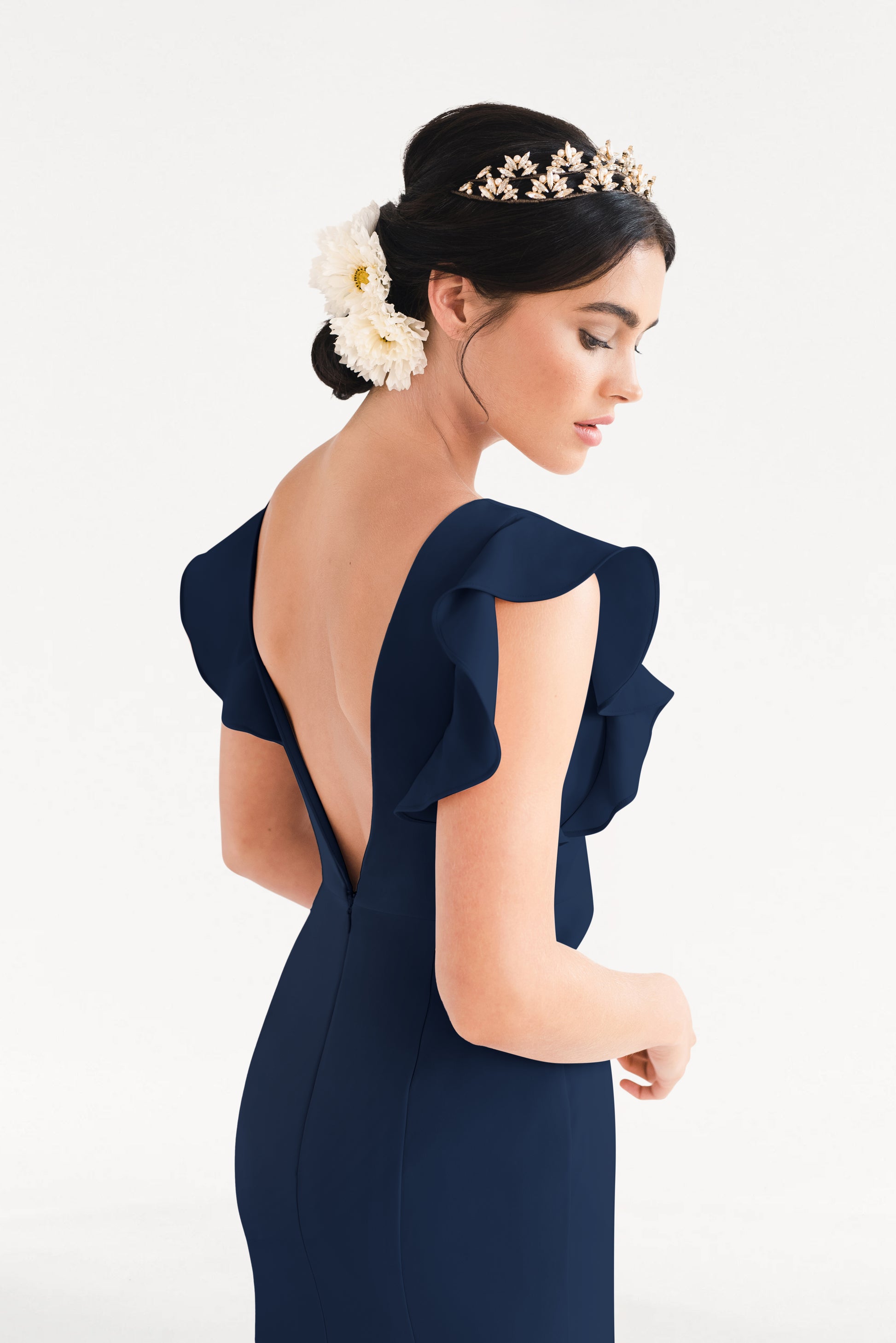 A woman in the TH&TH Cecelia Moss Crepe Bridesmaid Dress in Navy, featuring ruffled sleeves and a deep V-back, with a floral headpiece and large white flowers in her hair, looks down against a plain white background.