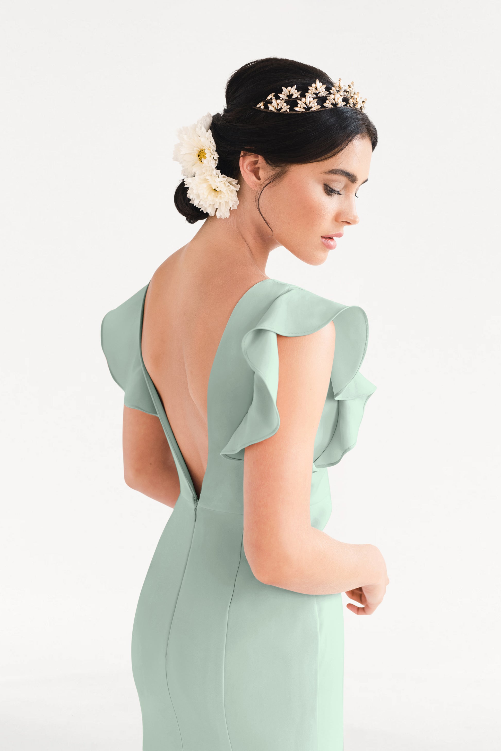 A woman with dark hair in an elegant updo, adorned with white flowers and a gold crown, wears the TH&TH Cecelia Moss Crepe Bridesmaid Dress in Sage Green featuring flutter sleeves and a deep V-back, standing against a plain white background.