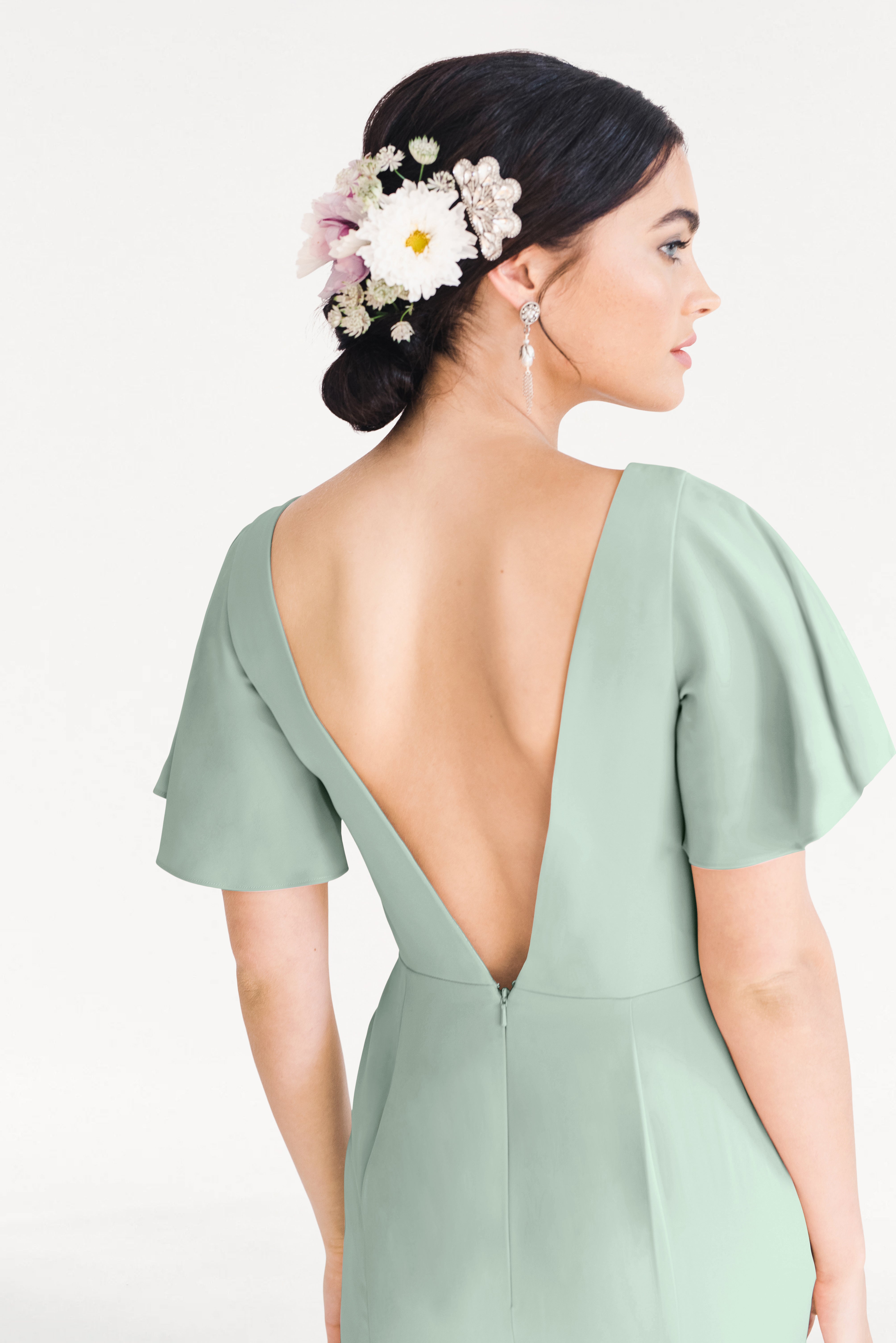 A woman wears the TH&TH Celeste Moss Crepe Bridesmaid Dress in Sage Green, a size-inclusive gown with a deep V-back and short sleeves. Her dark hair is in a low bun with white and pink flowers, complemented by dangling earrings against a plain background.