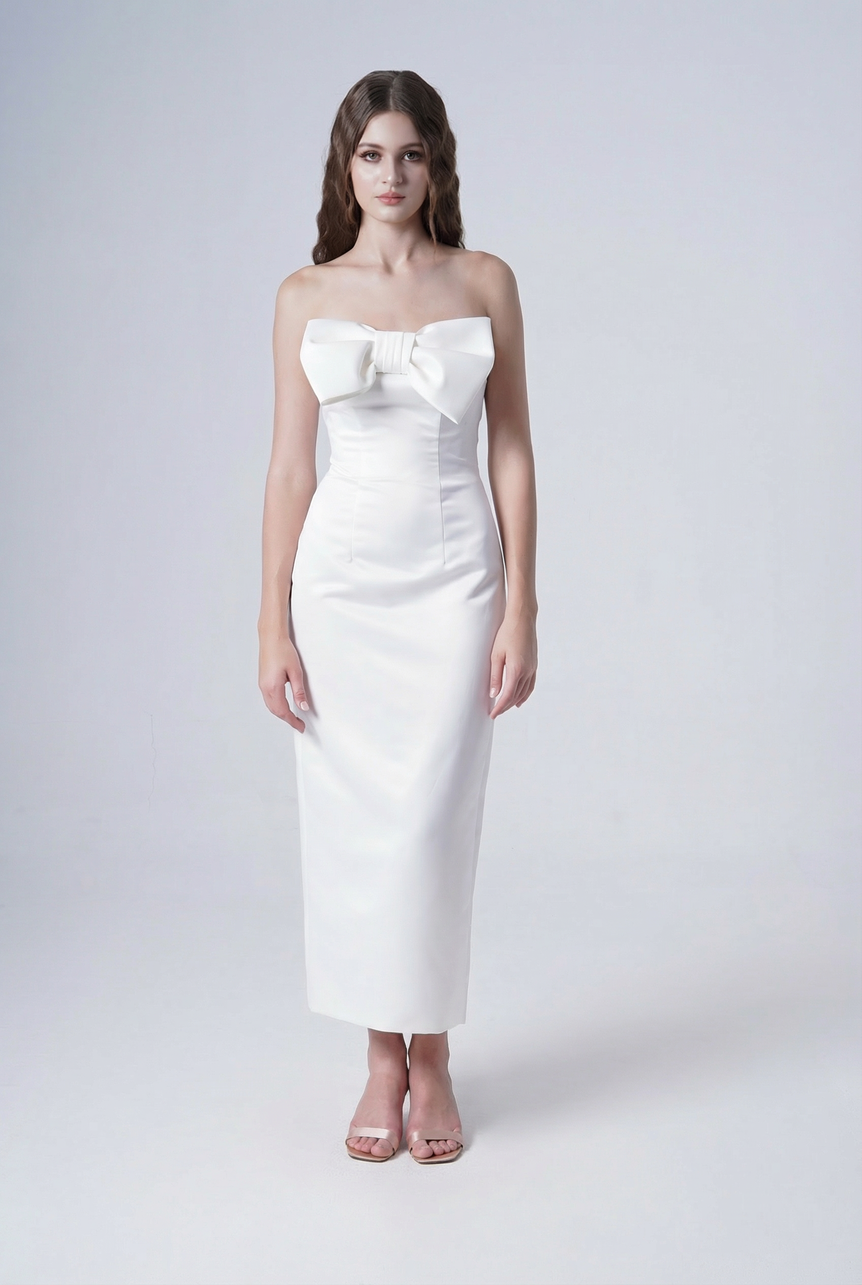 A woman stands against a plain background wearing the TH&TH Ivy Bridal Dress in Ivory—a white, strapless column gown with a large chest bow—paired with open-toed heels.
